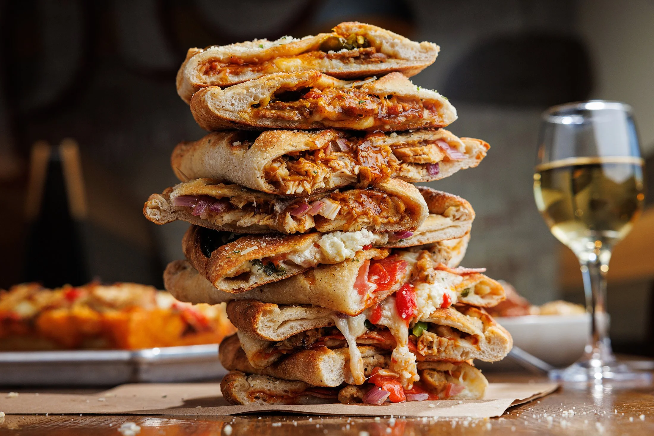 Stack of assorted calzones with melted cheese, meats, and vegetables on a wooden surface next to a glass of white wine.