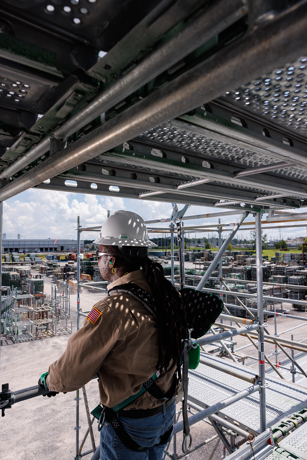 A construction worker with a safety helmet and harness standing on scaffolding at an outdoor construction site, with many construction materials and equipment in the background.