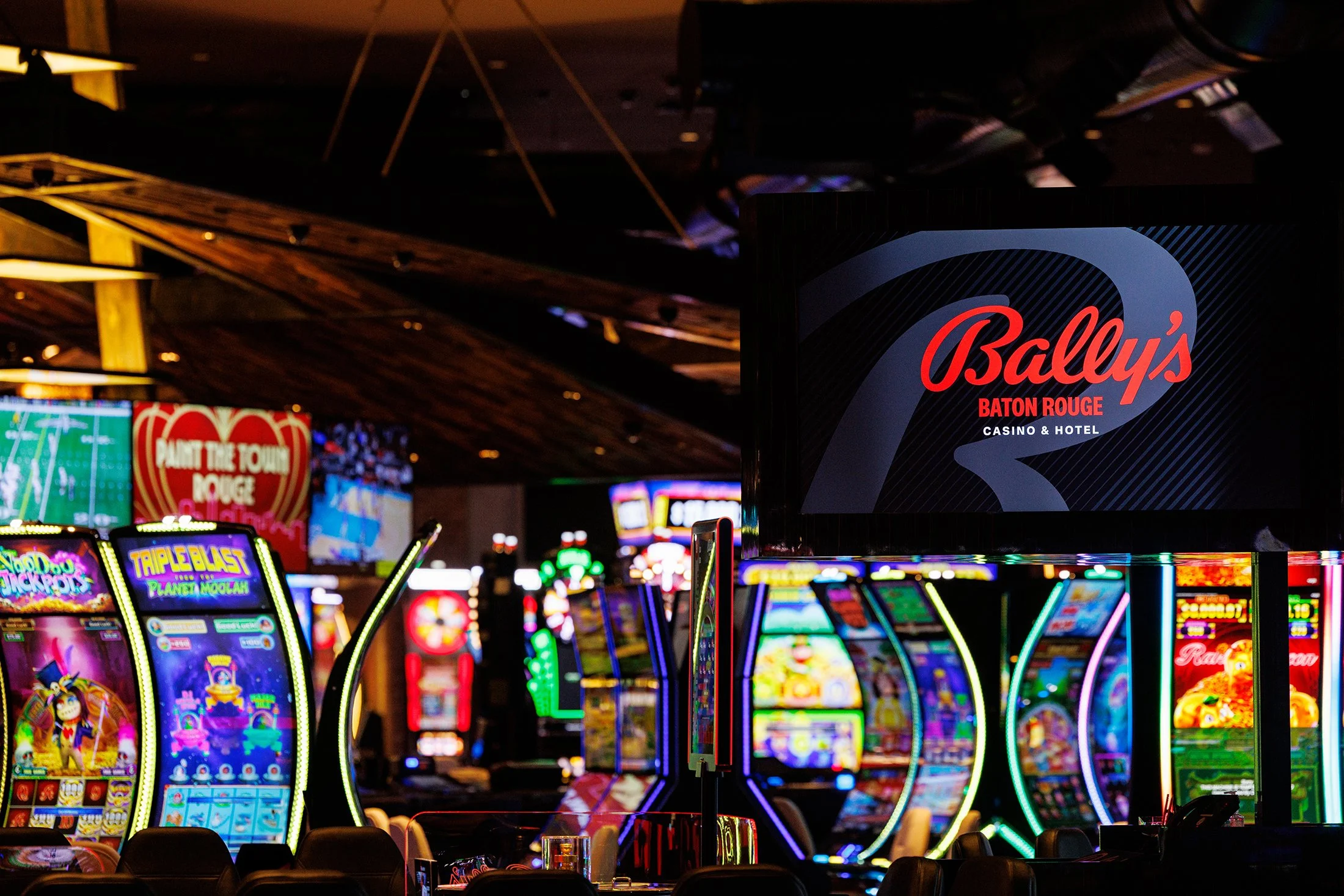 Inside a casino with bright, colorful slot machines and a large sign for Bally's Baton Rouge Casino & Hotel.
