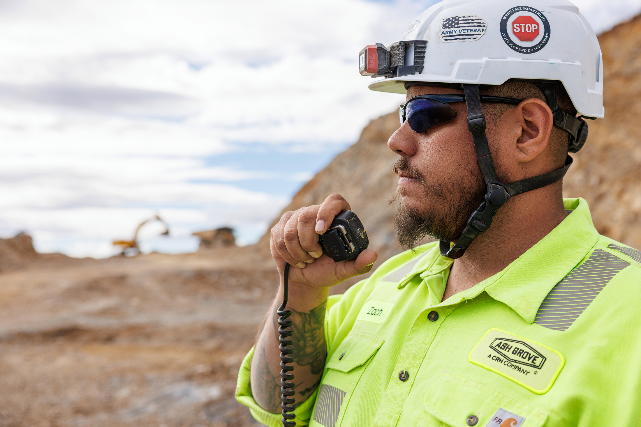 A construction worker wearing a white safety helmet with stickers, sunglasses, and a high-visibility yellow shirt, holding a walkie-talkie at a worksite with dirt and machinery in the background.