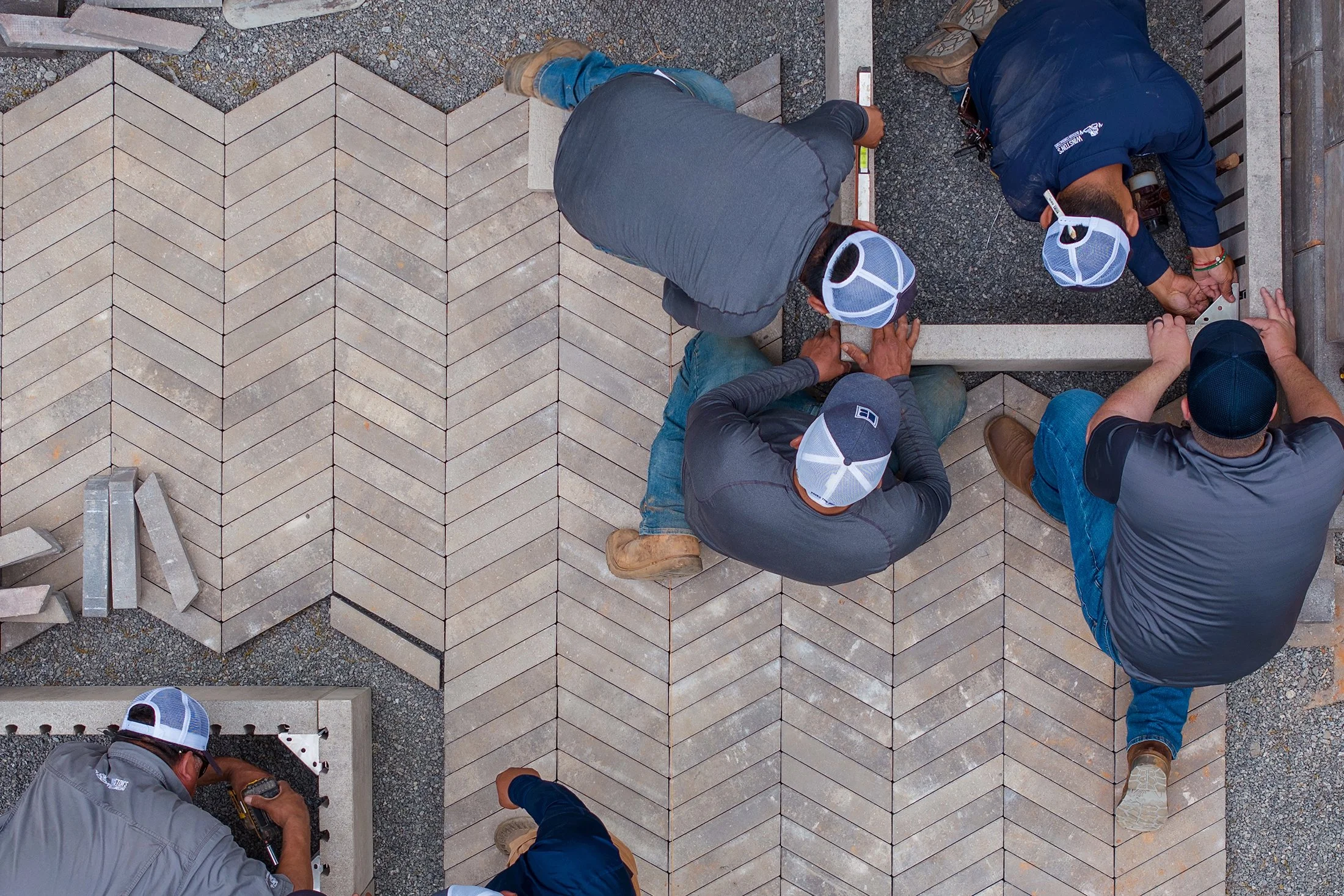 Six construction workers laying rectangular paving stones in an outdoor area, working together on the ground.