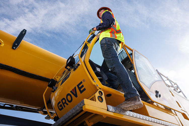 Construction worker wearing a safety vest and helmet, standing in an excavator bucket, working on a large yellow pipeline at a construction site.