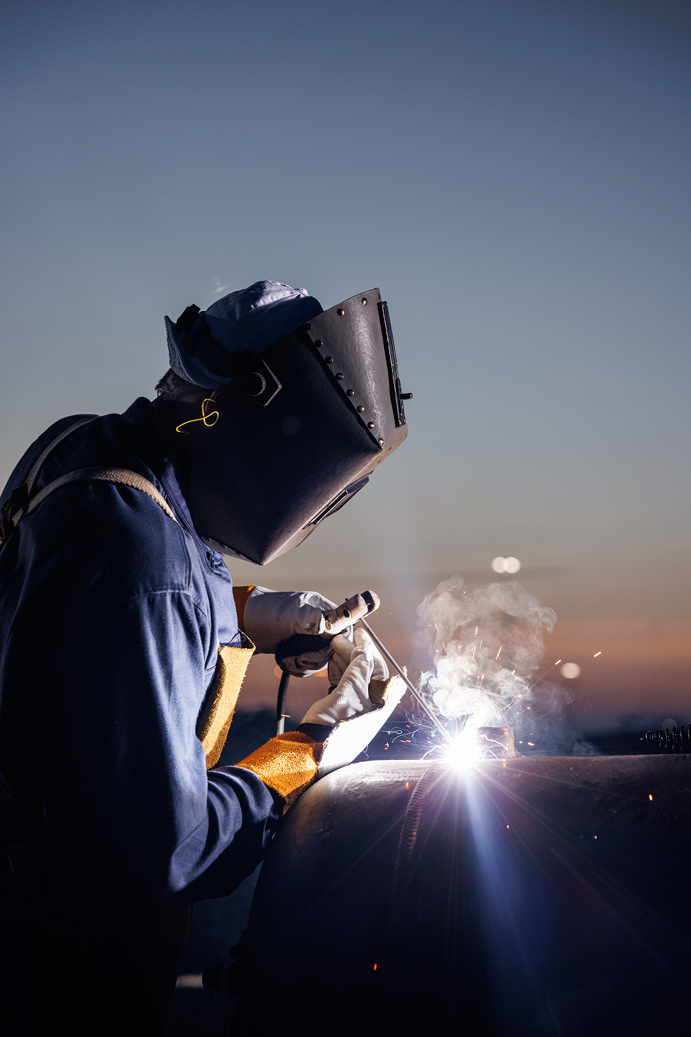 A welder wearing protective gear, including a welding helmet, gloves, and a work jacket, is welding metal outdoors at sunset, with sparks and smoke visible.