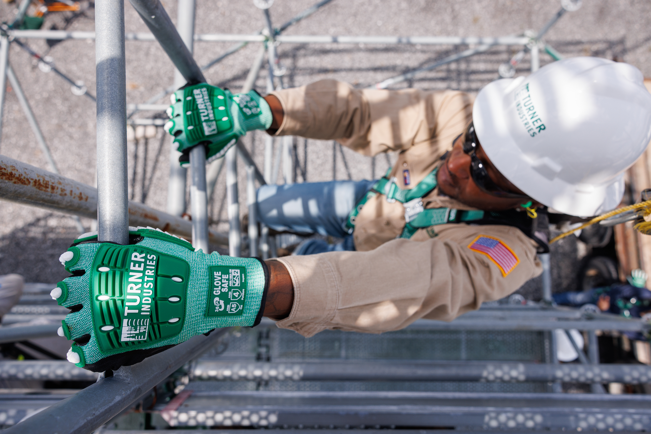 A construction worker wearing tan uniform, white safety helmet, and green gloves is working on scaffolding.
