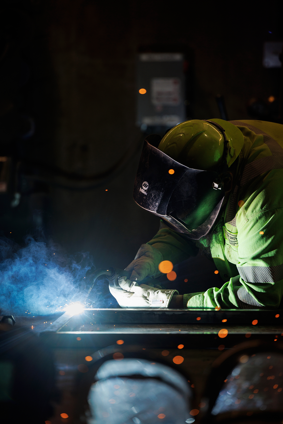 Welding worker wearing a yellow helmet and safety gear welding metal with sparks and smoke in a dark environment.