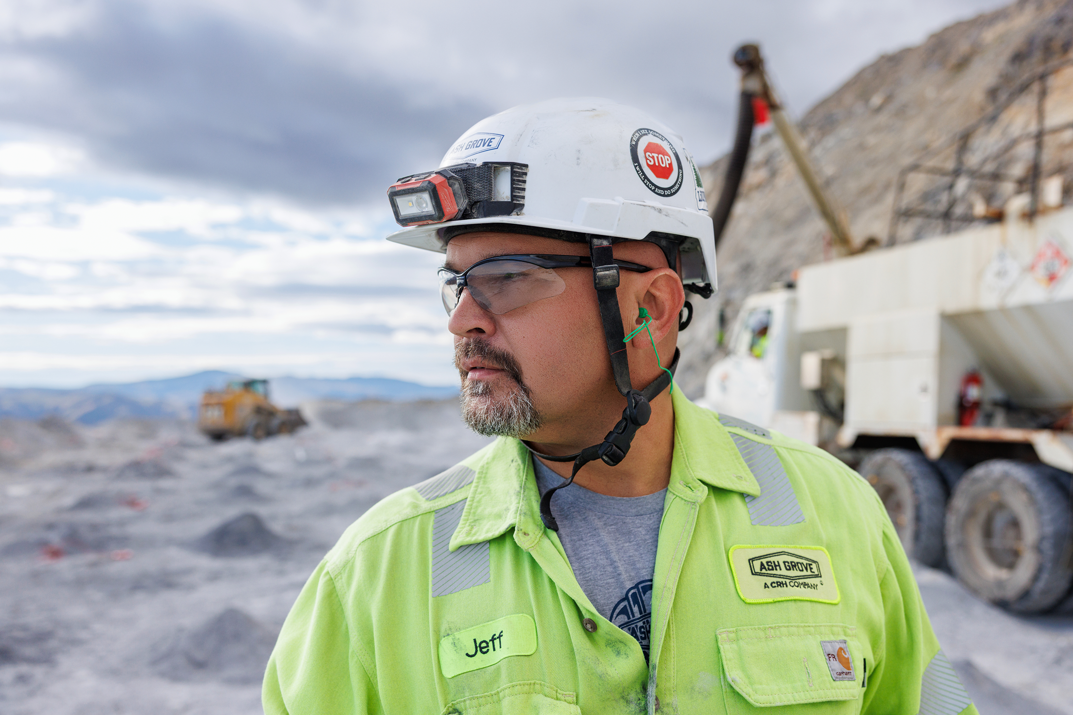 A male mining worker wearing safety goggles, a white hard hat with a light, and a neon yellow safety jacket with the name tag 'Jeff,' standing in a mining or construction site with trucks in the background and mountainous terrain under a cloudy sky.