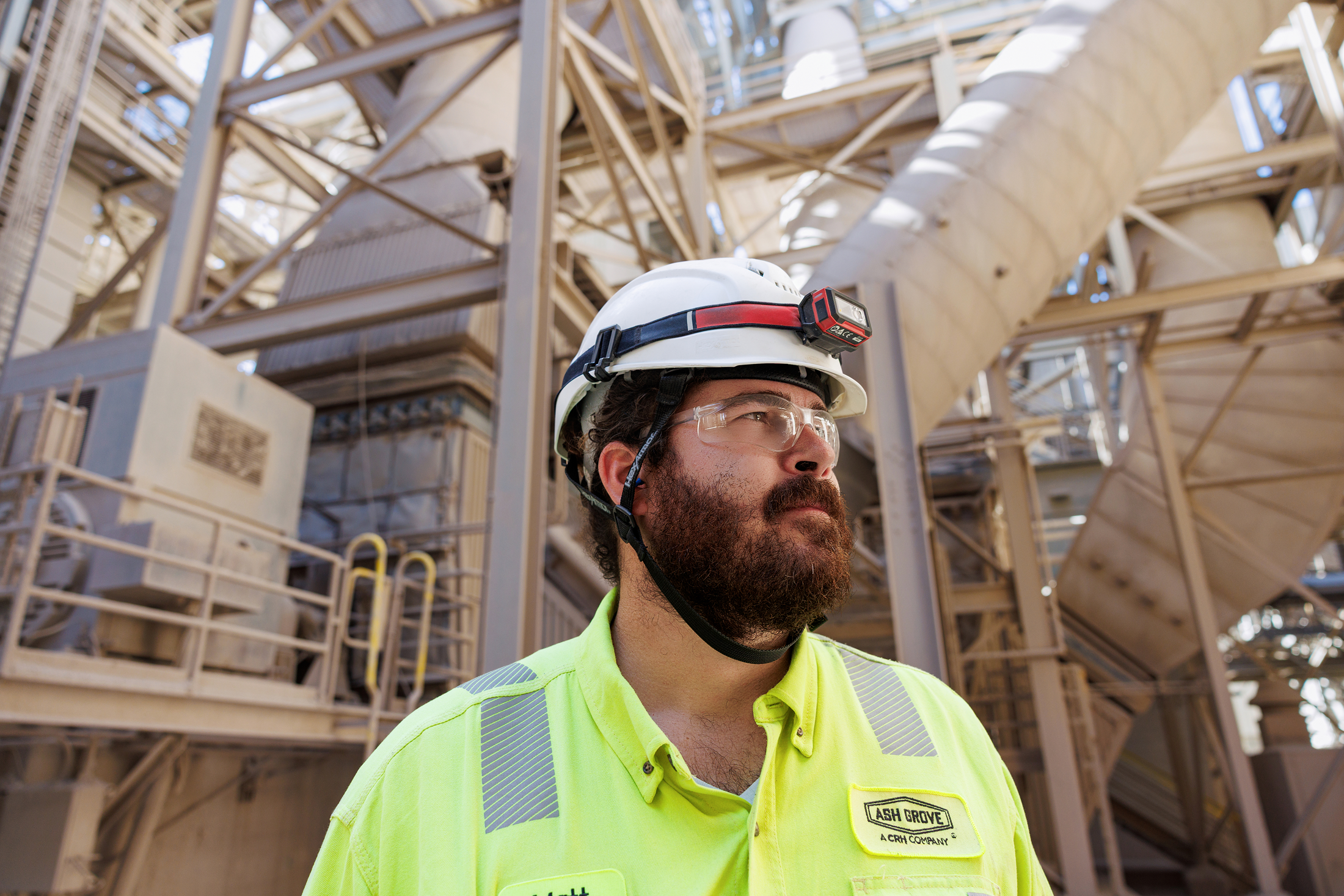 A construction worker wearing a yellow high-visibility shirt, safety glasses, a white hard hat with a headlamp, standing in front of industrial equipment and structure at a construction site.