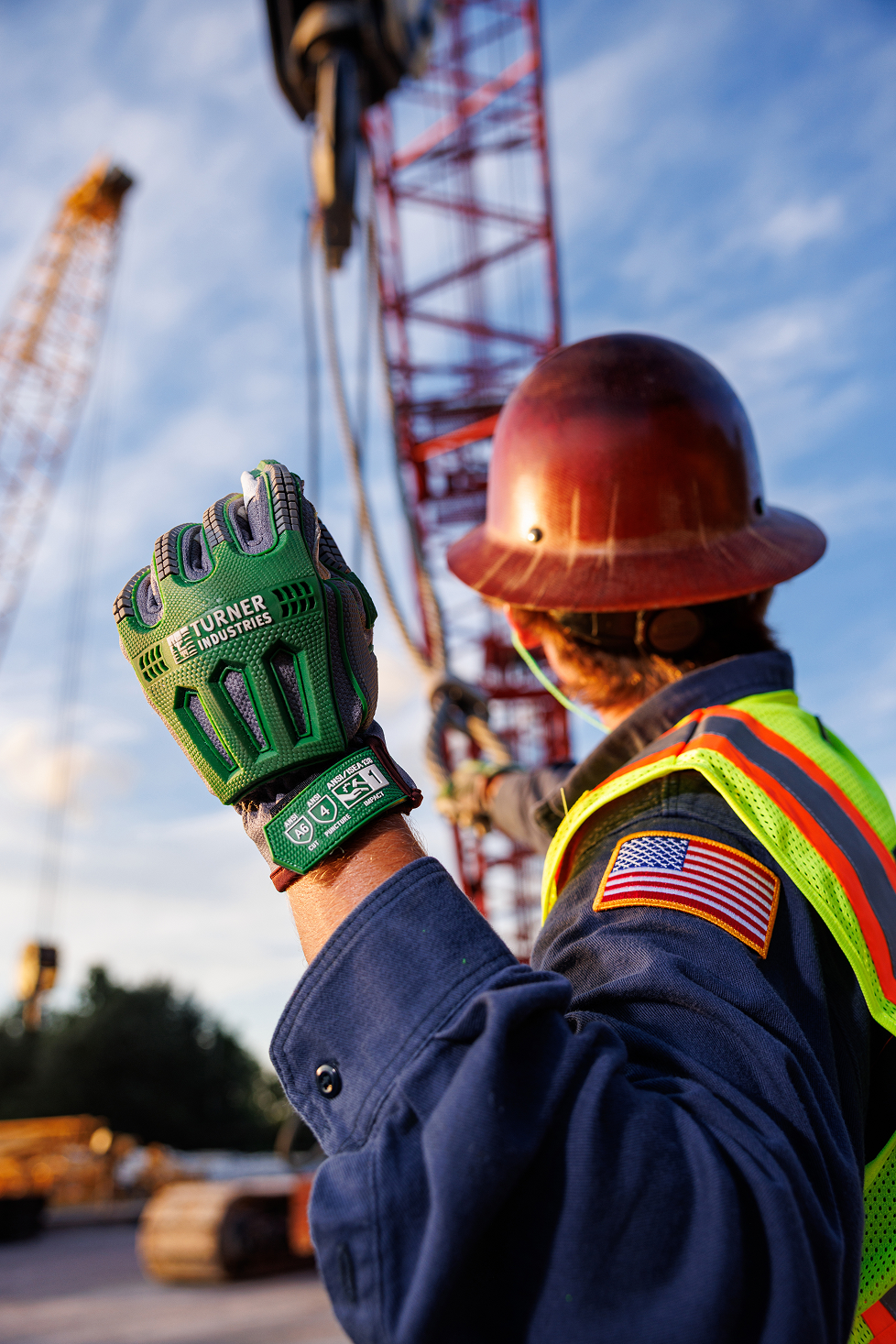 A construction worker wearing a brown helmet, safety vest, and a US flag patch on the sleeve, is working at a construction site next to a large red crane, with the worker's hand raised wearing a green heavy-duty glove, during late afternoon or early evening.