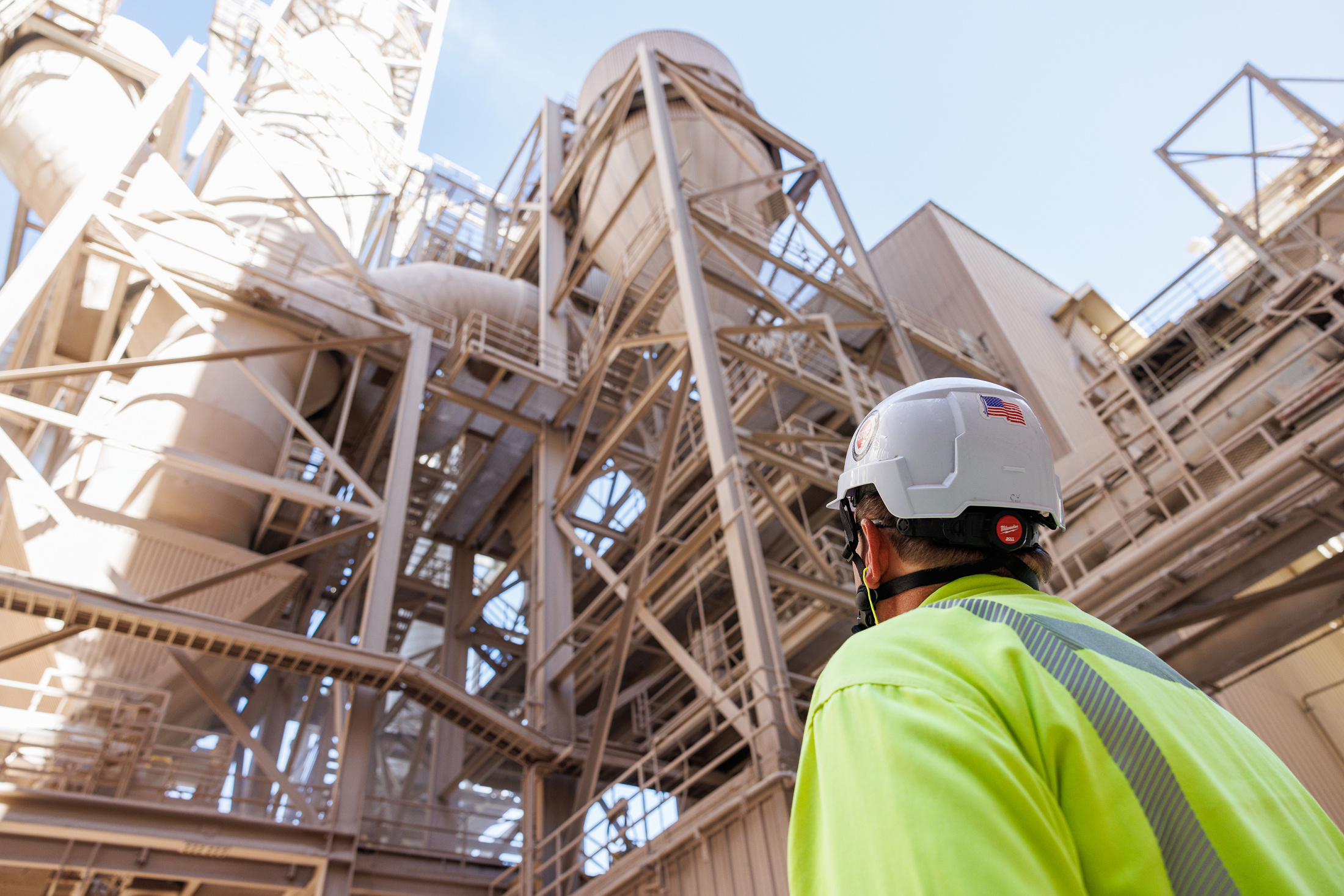 A worker wearing a yellow safety jacket and a white safety helmet overlooking an industrial plant with metal structures and pipelines.
