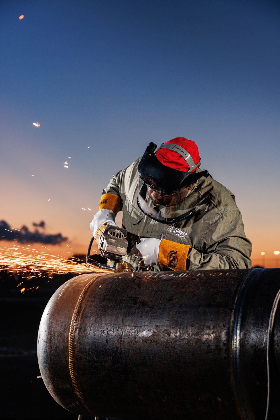 A worker in protective gear uses a grinder to cut or smooth a large metal pipe outdoors at sunset, with sparks flying.