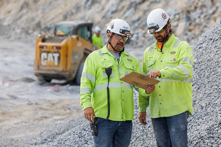 Two construction workers wearing bright green safety jackets and white helmets review a clipboard on a rocky construction site with heavy equipment in the background.