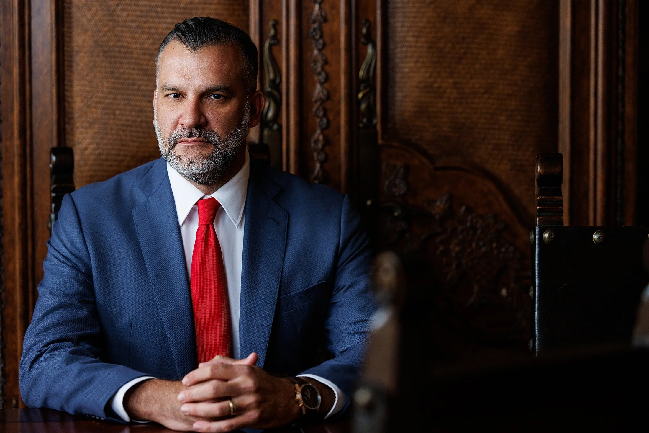 A man with dark hair and a beard, dressed in a blue suit, white shirt, and red tie, sitting indoors at a wooden table with ornate wooden paneling behind him.