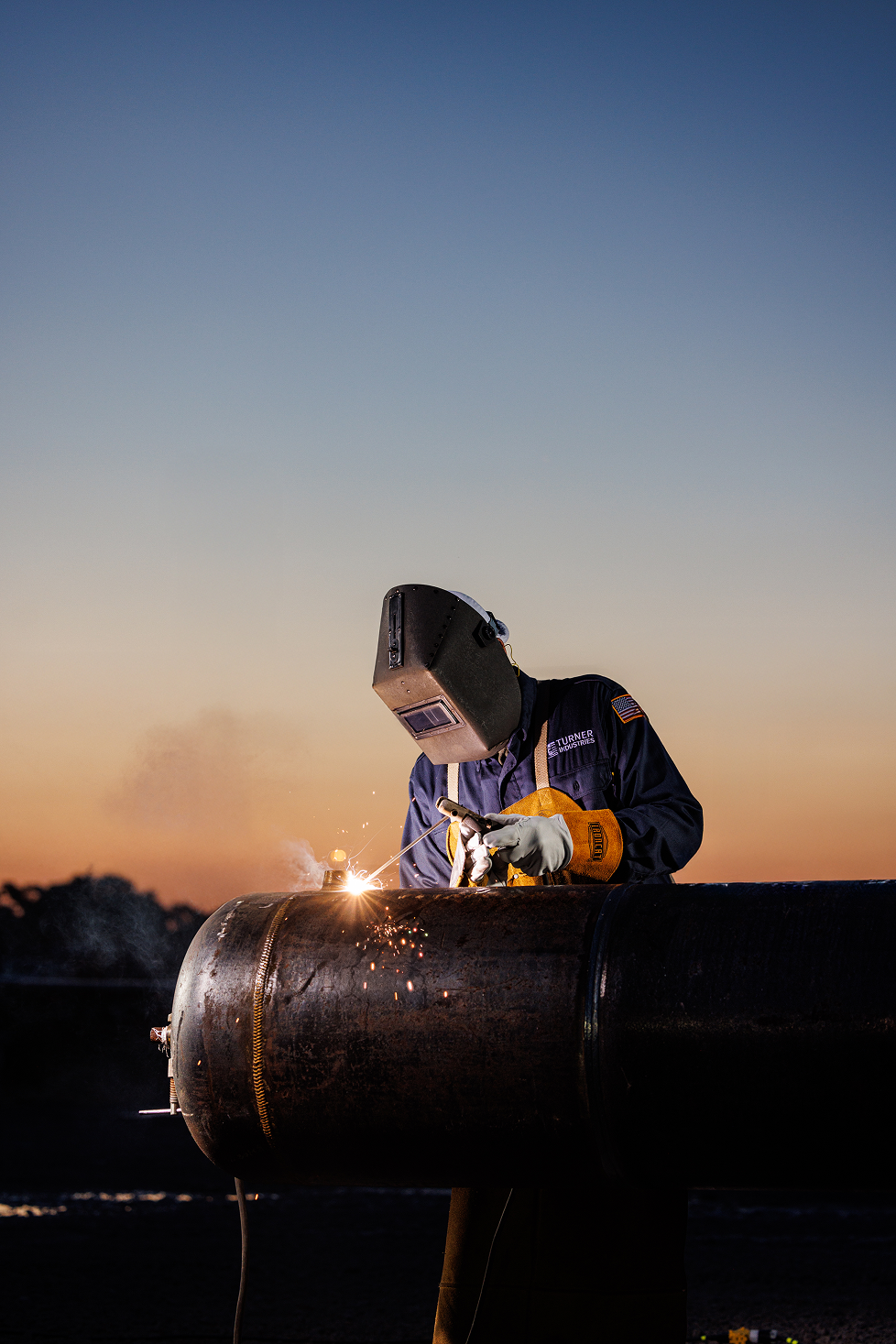 A welder wearing protective gear, including a helmet and gloves, welding a large metal pipe outdoors at sunset.