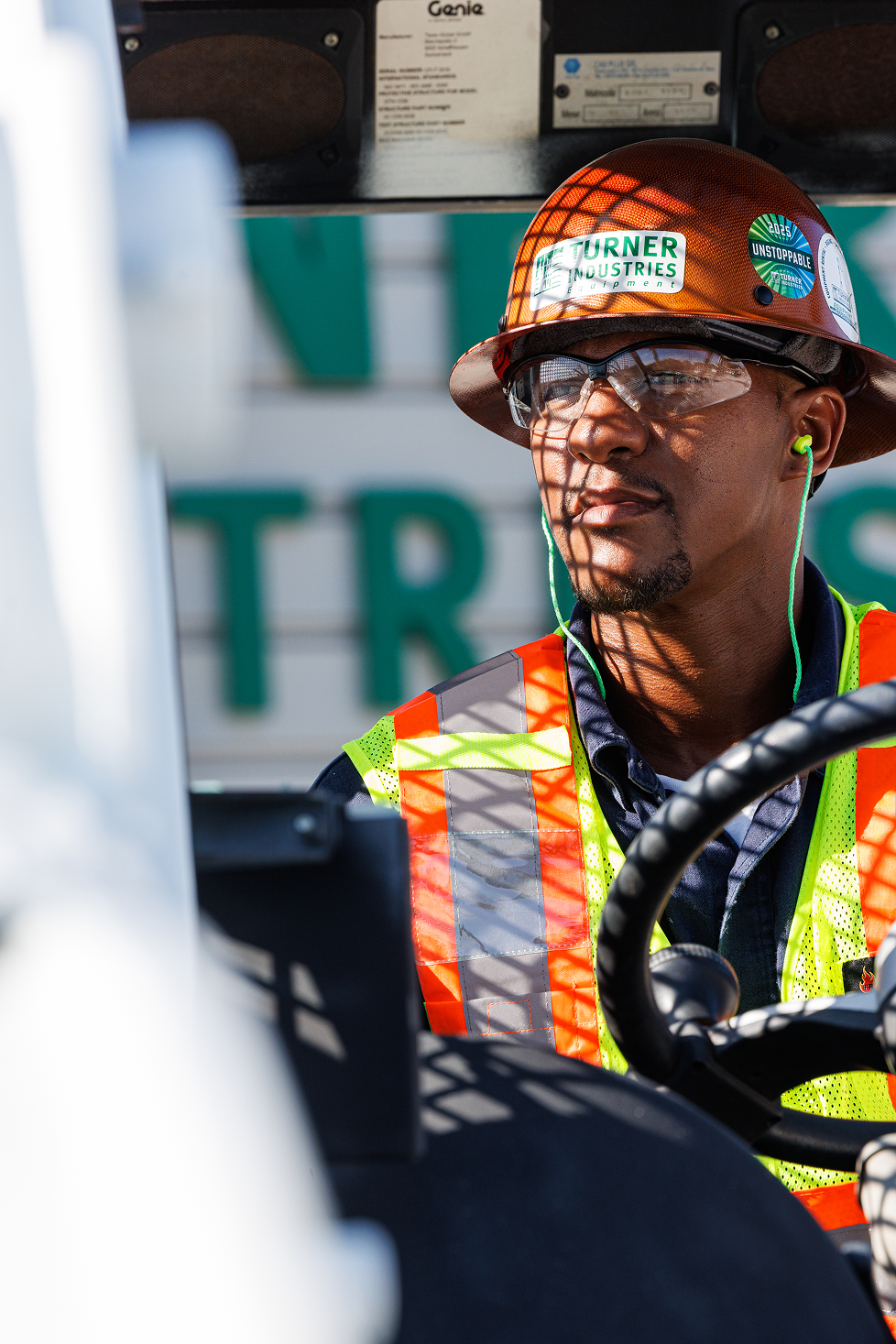 A construction worker wearing a brown safety helmet with stickers, clear safety glasses, a high-visibility orange and yellow vest, and green earplugs, sitting at a construction site.