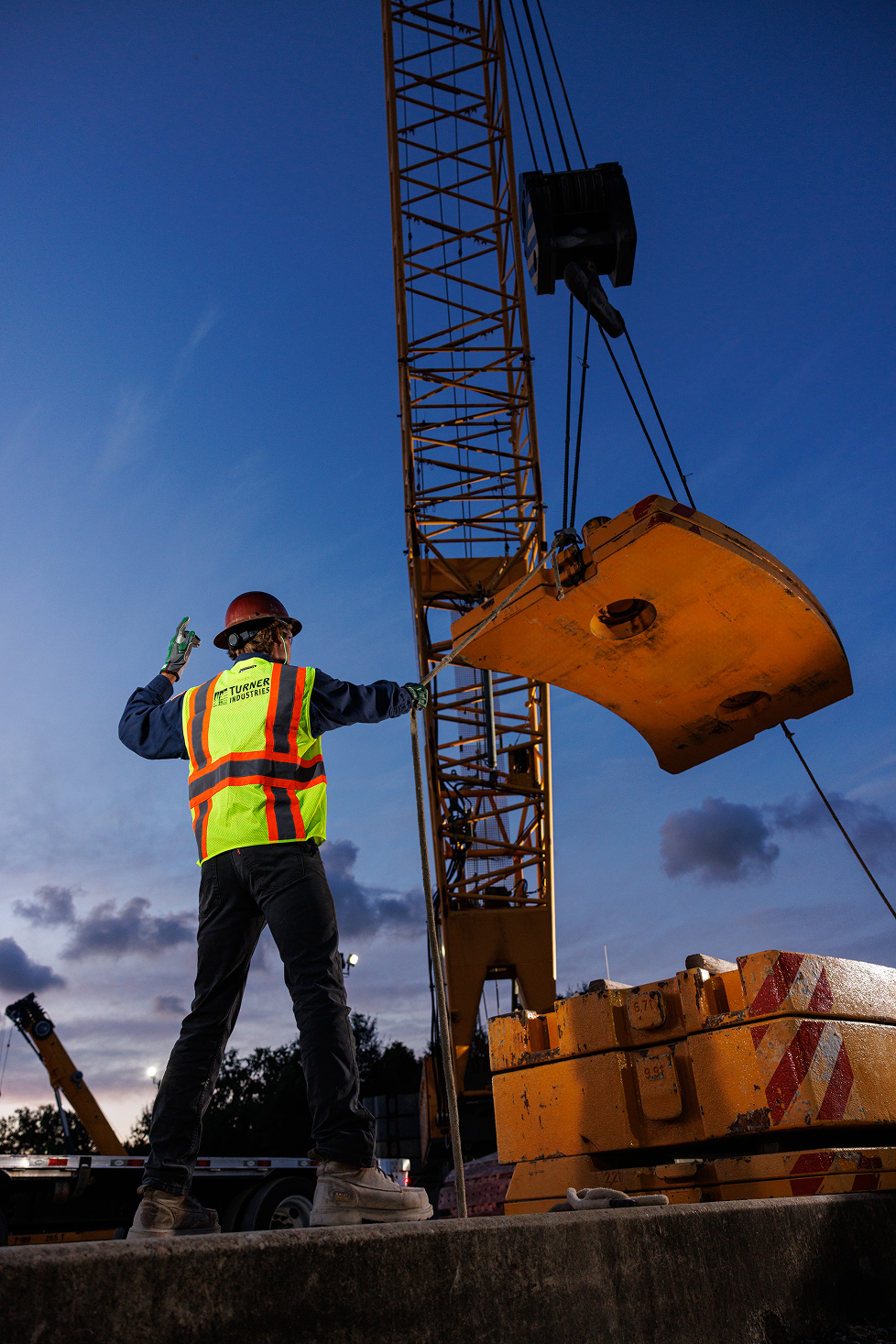 Construction worker wearing safety gear adjusting a crane at sunset.