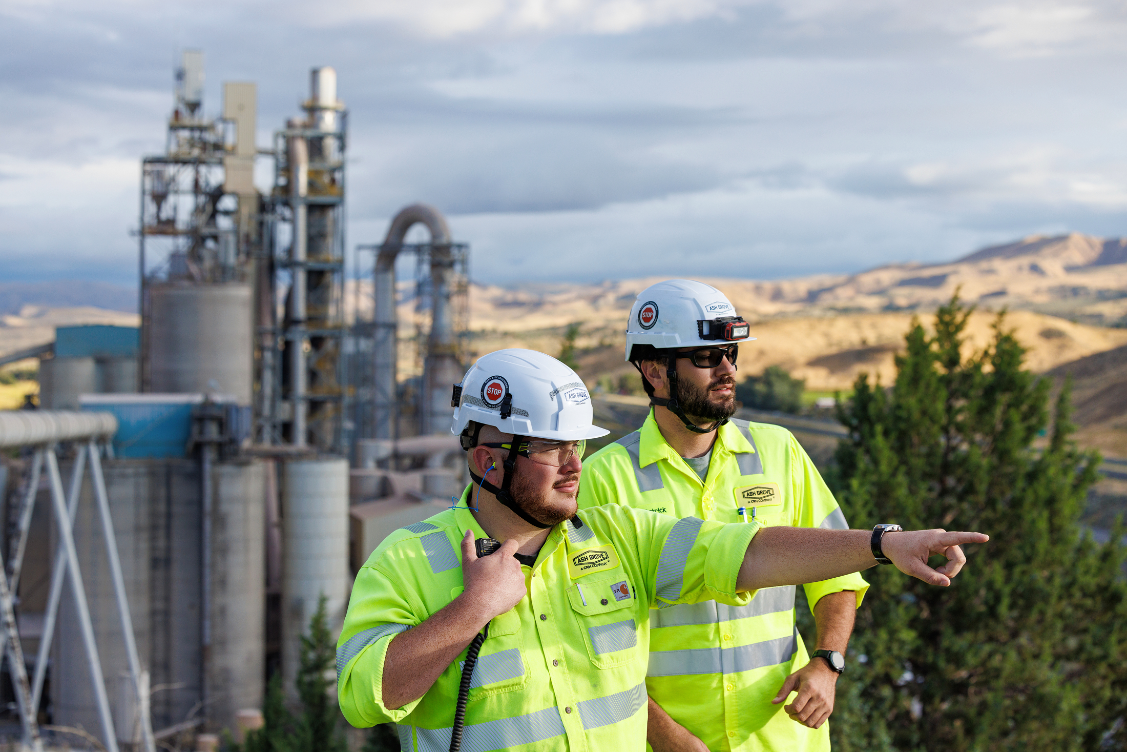 Two male workers wearing safety gear, including helmets and high-visibility shirts, standing outdoors at an industrial site. One is pointing towards the distance.