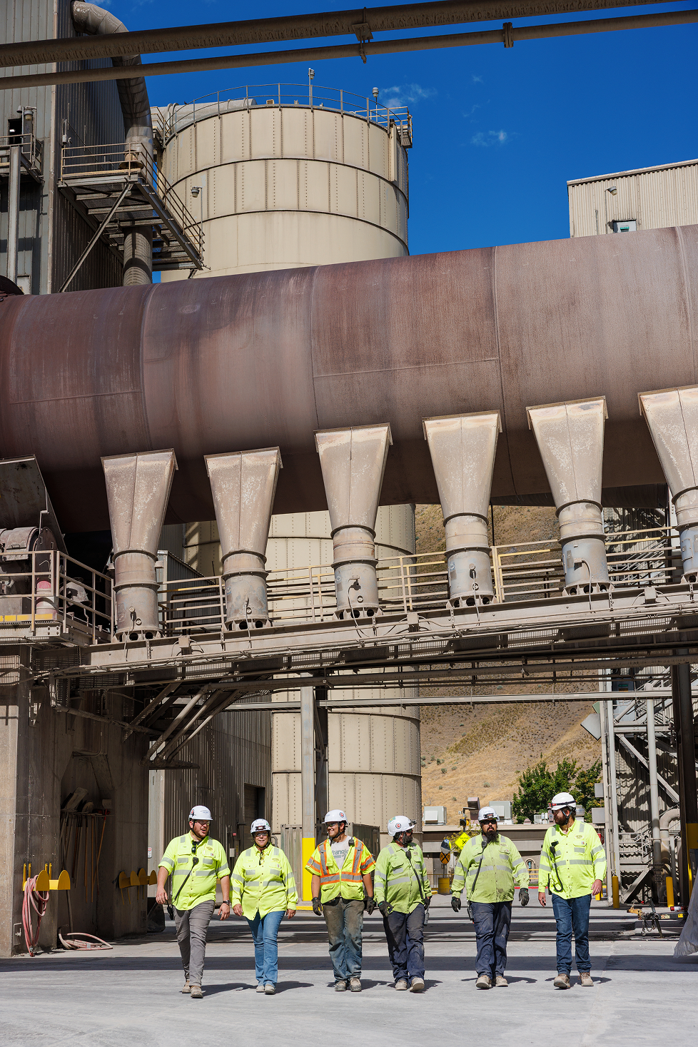 Six workers wearing safety helmets and reflective vests walk in an industrial factory with large pipes and tanks behind them.