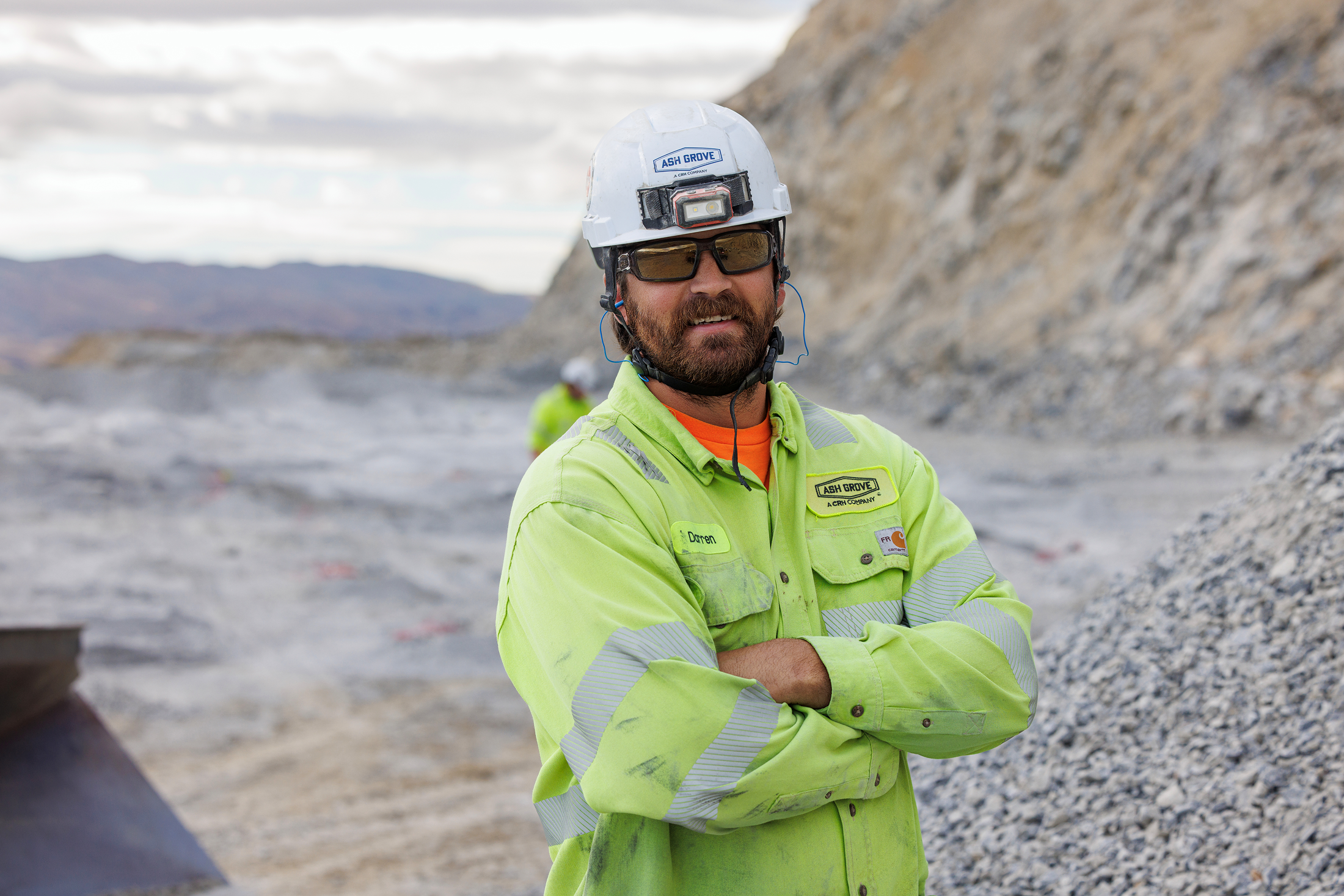 A man wearing a high-visibility safety shirt, helmet, sunglasses, and ear protection stands outdoors at a construction or mining site with rocky terrain and mountains in the background.