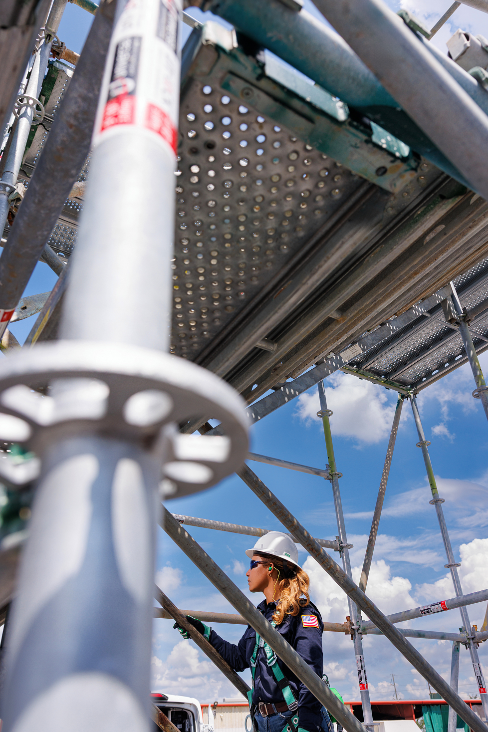 A female construction worker wearing a white hard hat, sunglasses, and ear protection working underneath scaffolding on a construction site.