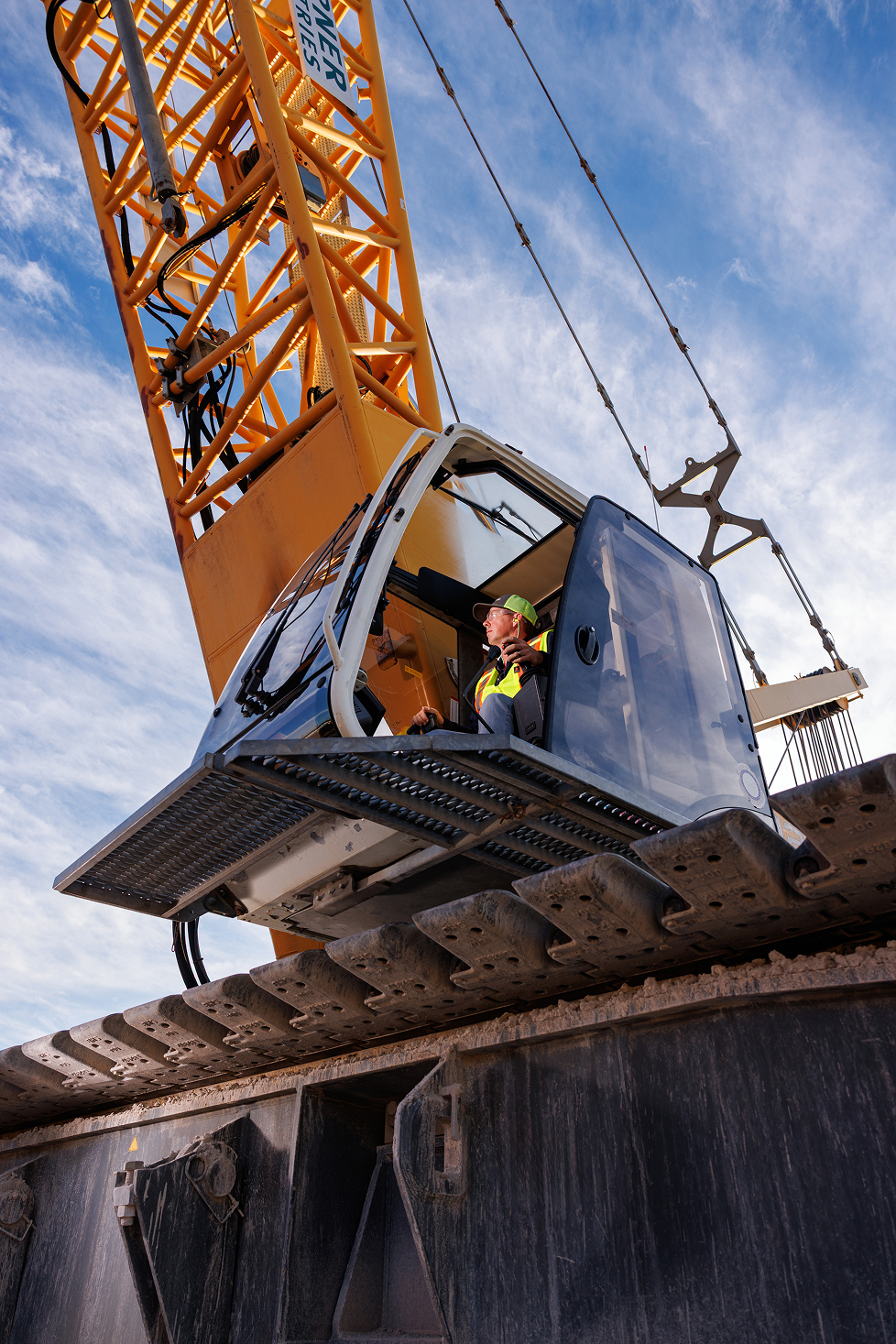 Close-up of a construction crane operator in a cab, wearing a yellow safety vest and cap, with a blue sky in the background.
