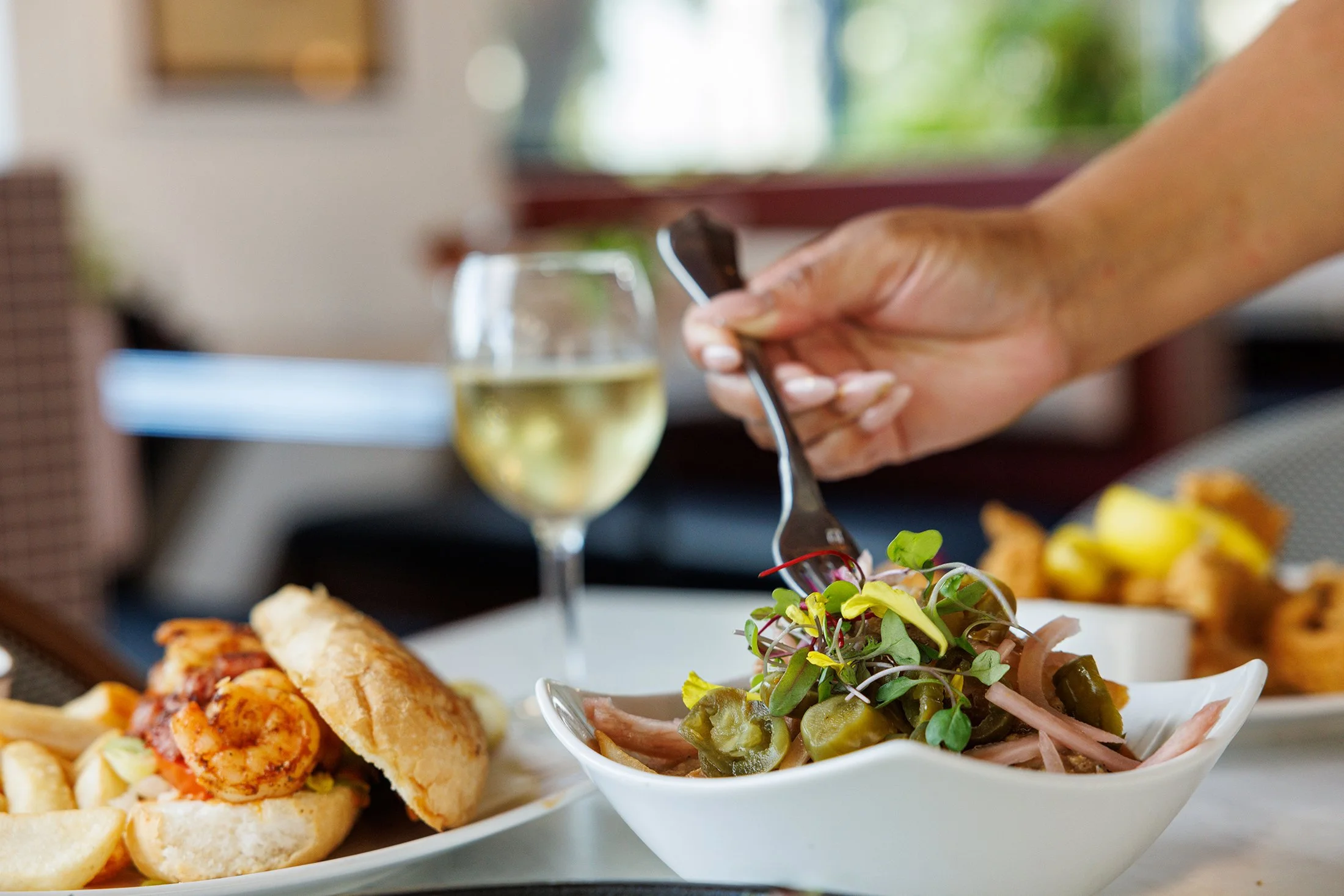 Person serving a mixed green salad with microgreens, vegetables, and toppings at a dining table, with a glass of white wine and plates of food nearby.