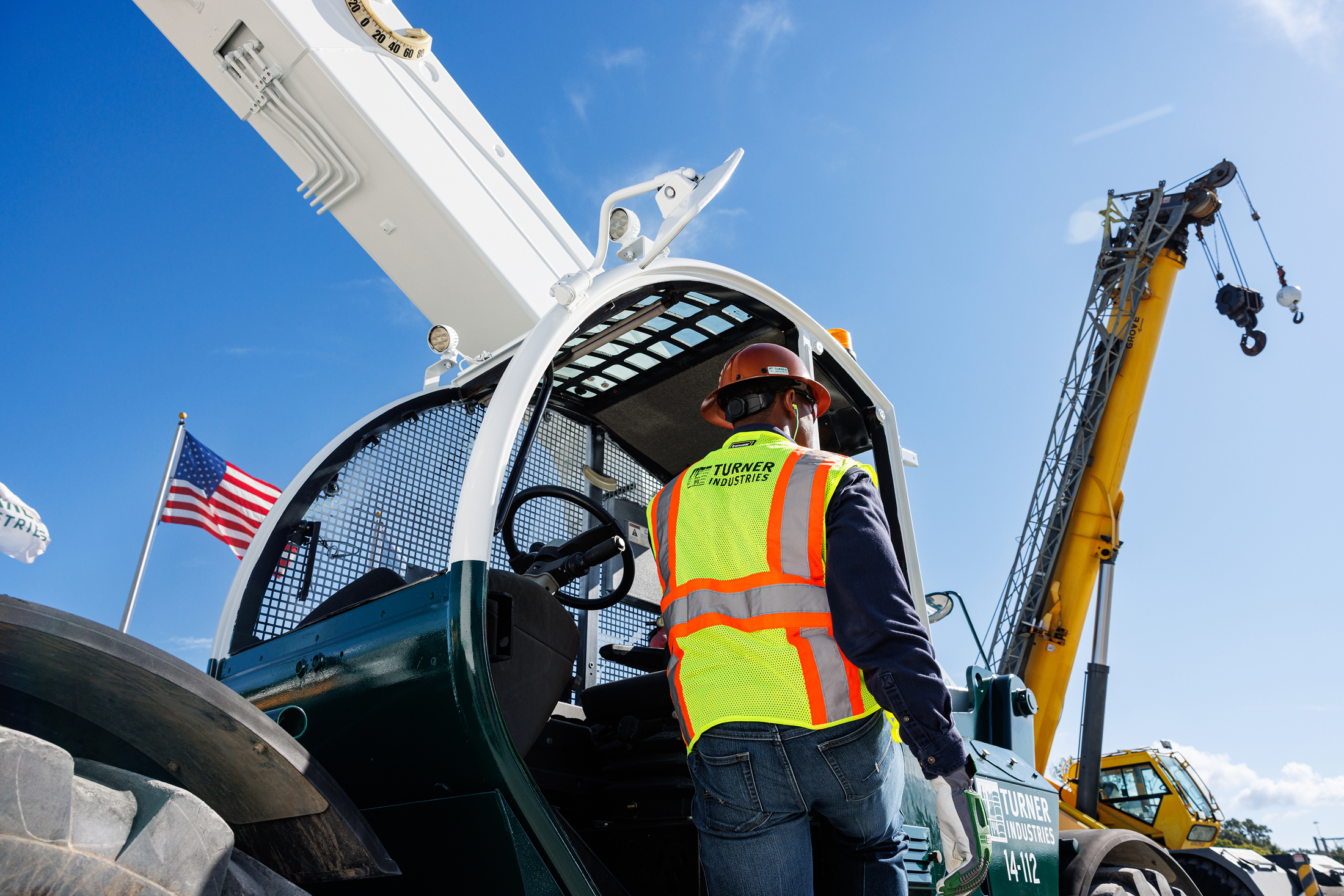 A construction worker wearing a yellow safety vest and a hard hat standing next to a crane truck on a bright, clear day with a blue sky, American flags, and yellow cranes in the background.