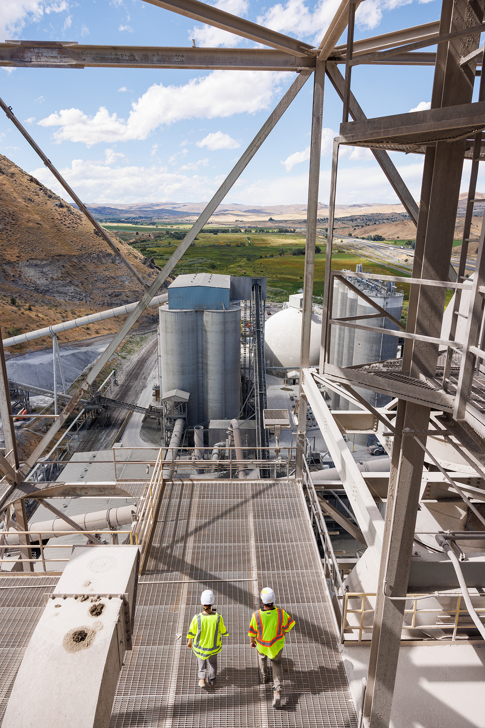 Two workers in safety vests and helmets walk on metal grating at an industrial plant, with large silos and pipes, overlooking a rural landscape with mountains and fields under a partly cloudy sky.