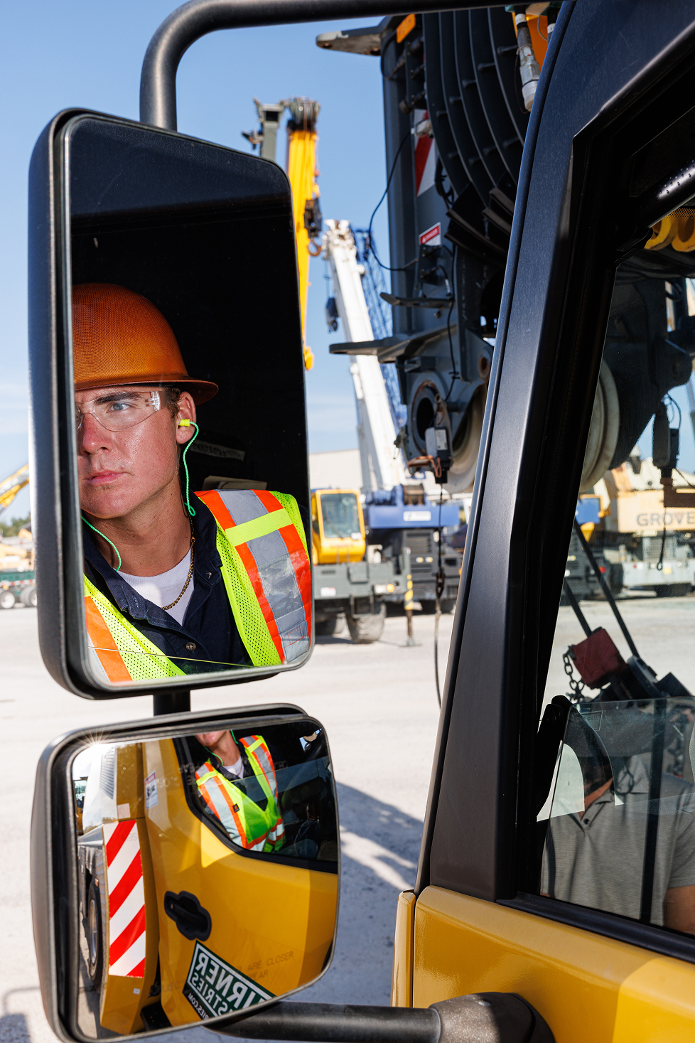 A male construction worker wearing a yellow hard hat and safety glasses looking into a side mirror of a construction vehicle. He is dressed in a high-visibility safety vest and is at a construction site with cranes and machinery in the background.