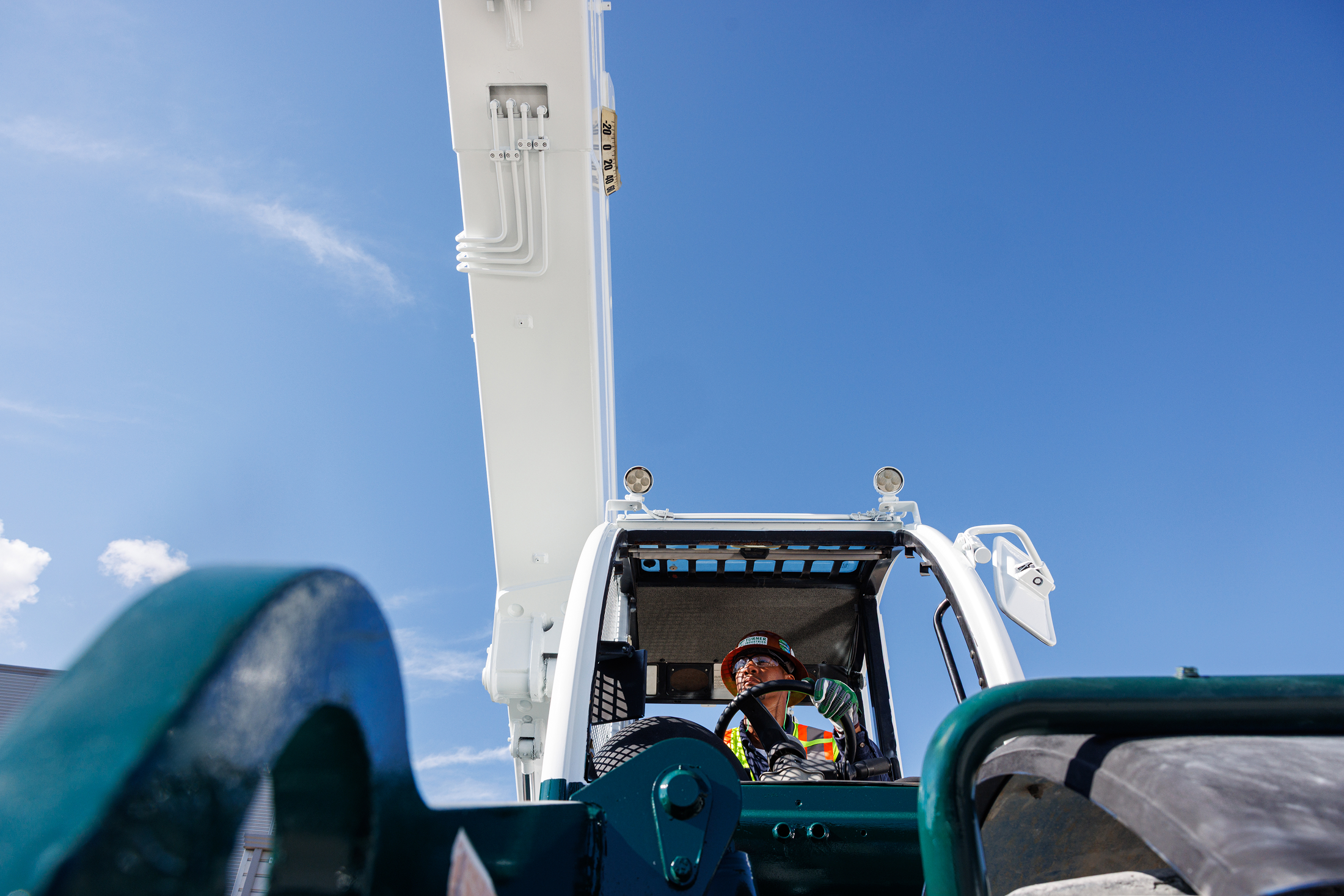 A construction worker wearing safety gear and a helmet sits inside a large construction vehicle with an extended arm against a clear blue sky.