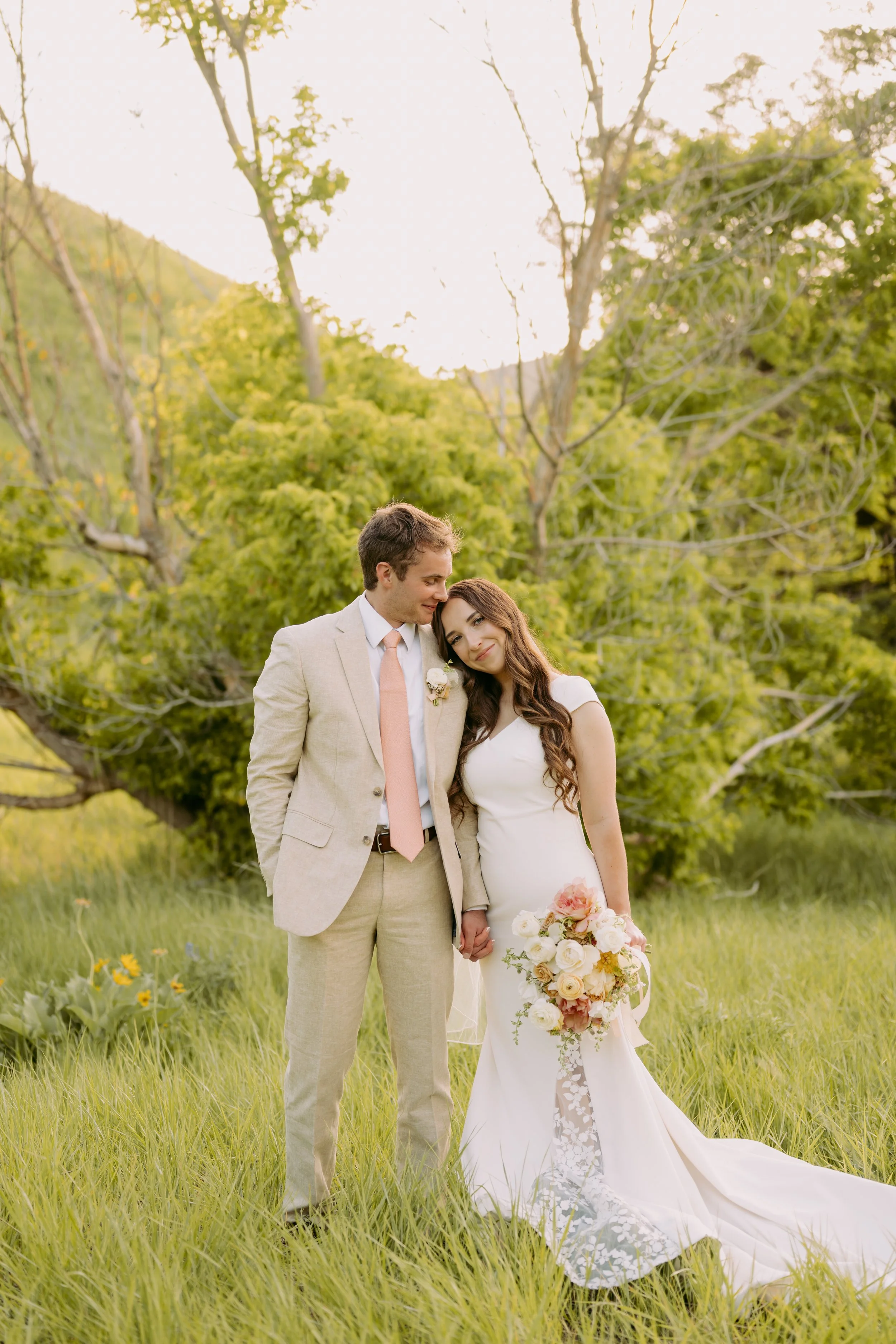 A bride and groom holding hands in a grassy field with green trees and hills in the background, during a wedding photoshoot.