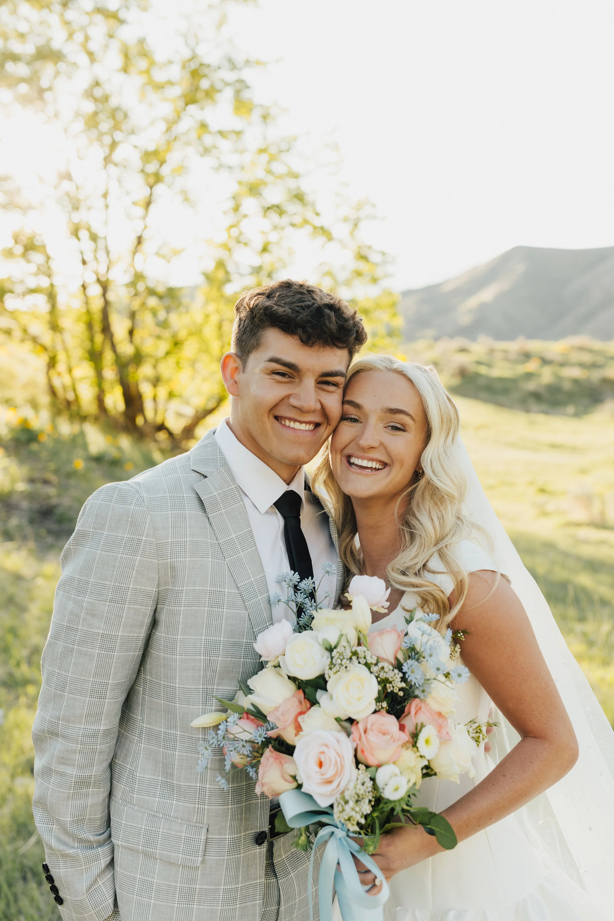 A newlywed couple, smiling and close together, outdoors with trees and mountains in the background. The bride holds a large bouquet of flowers.