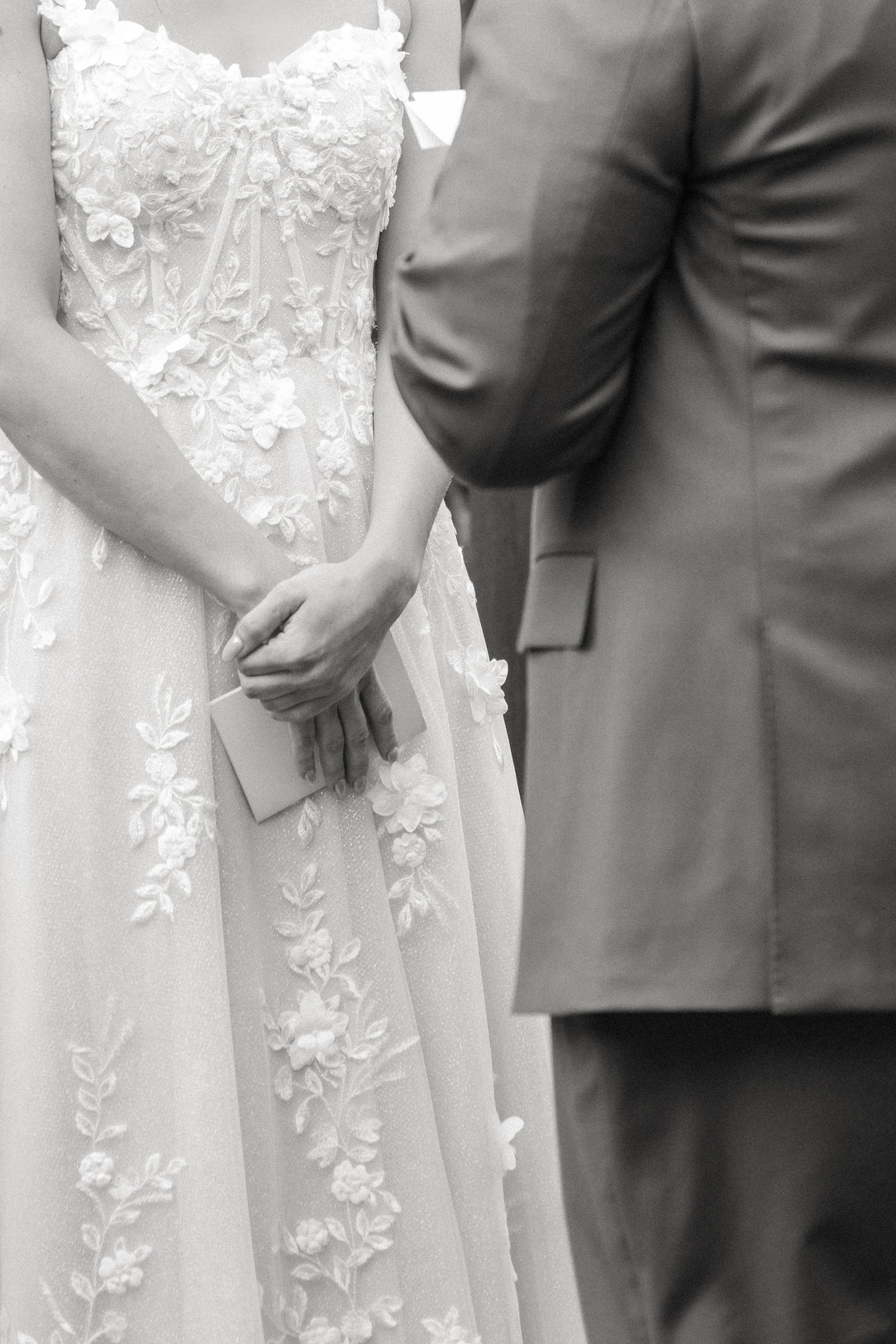 A close-up black-and-white photo of a couple holding hands during a wedding ceremony. The bride is wearing a detailed, floral lace wedding dress, and the groom is in a suit.
