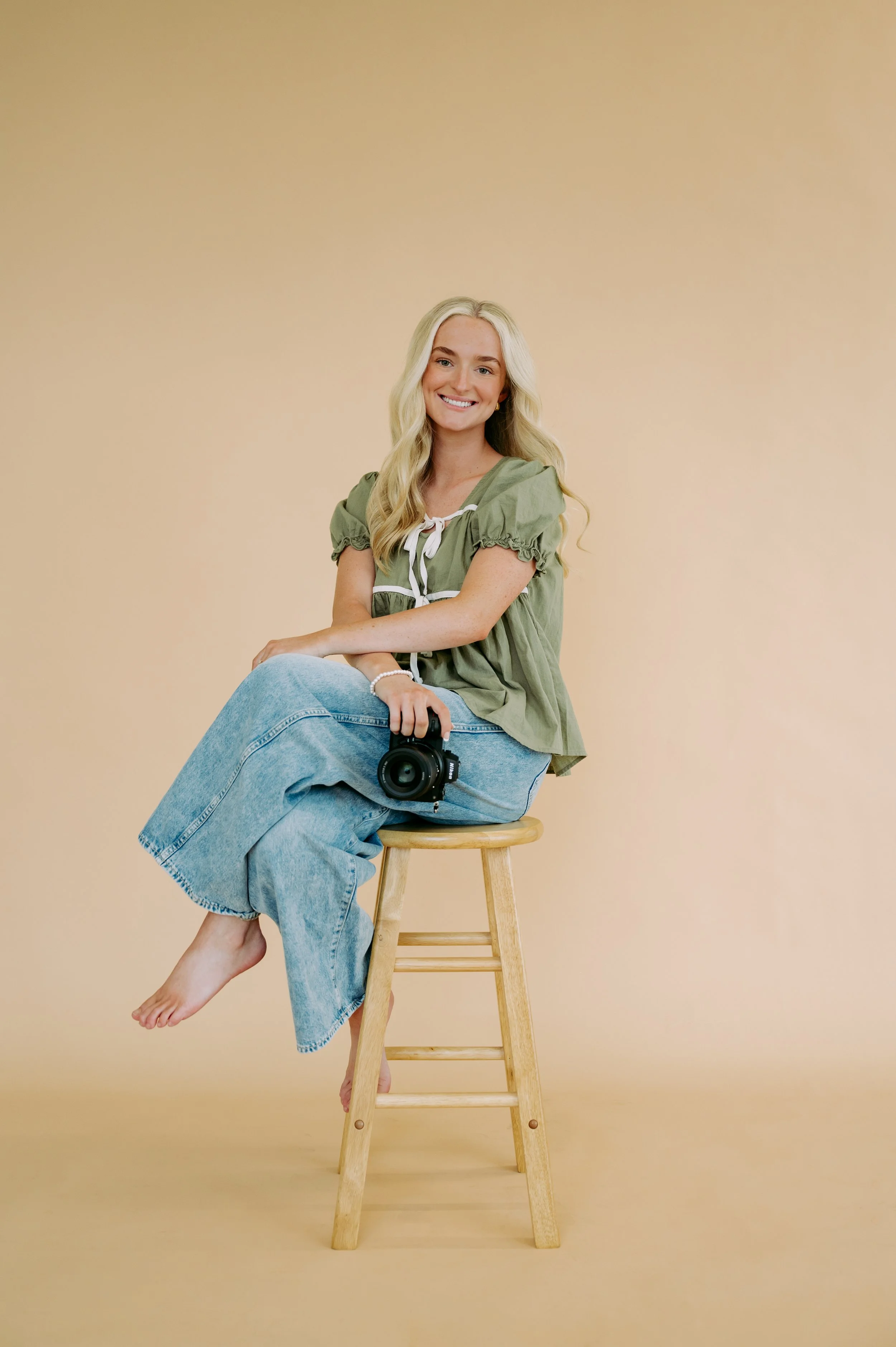 A young woman with long blonde hair, wearing a green top and light blue jeans, is sitting on a wooden stool, holding a camera, and smiling at the camera against a beige background.