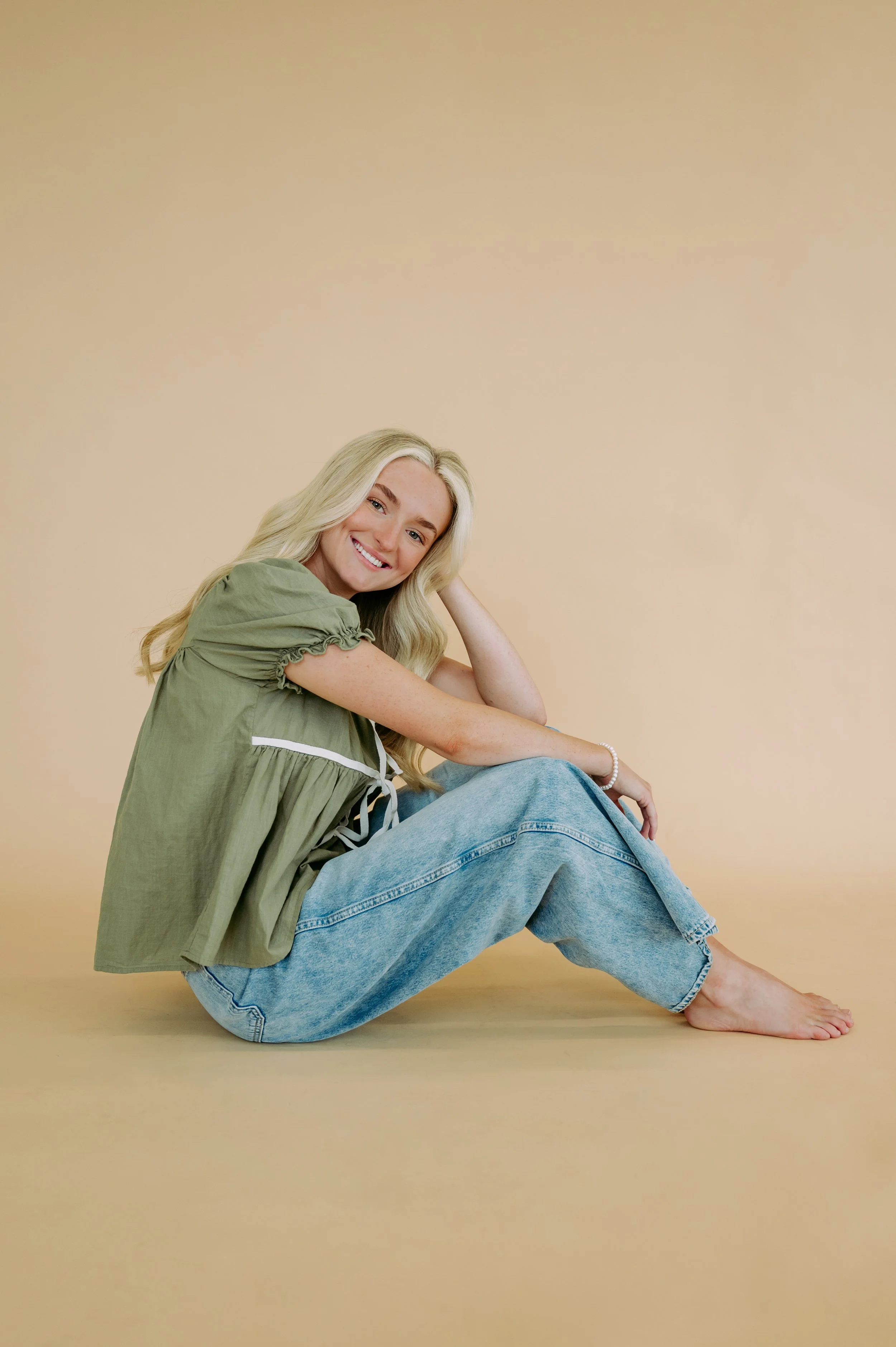A woman with long blonde hair, wearing a green top with puffed sleeves and light blue jeans, sitting on the floor smiling at the camera against a beige background.