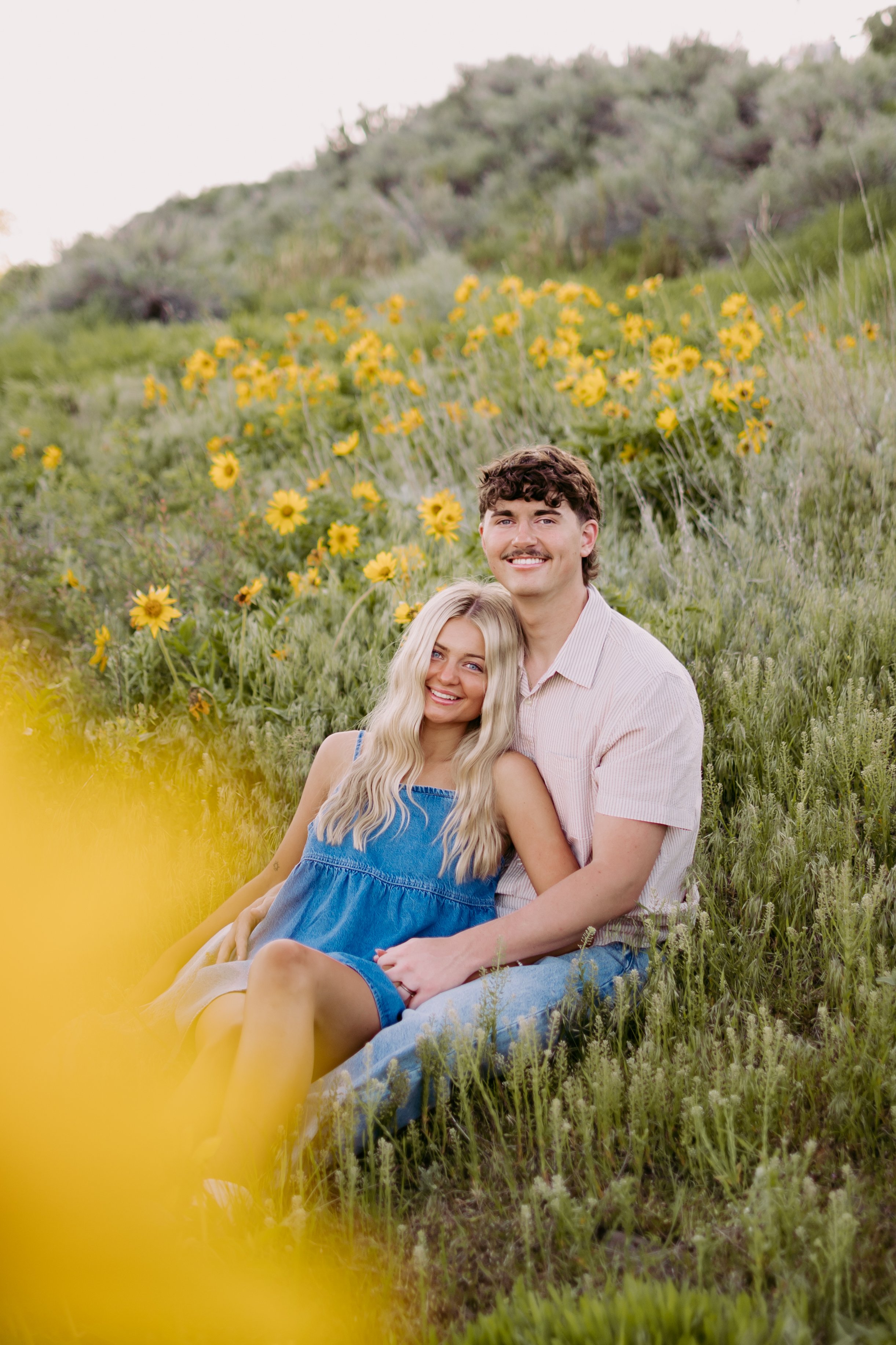A young couple sitting on grass in a field with yellow flowers, smiling at the camera.