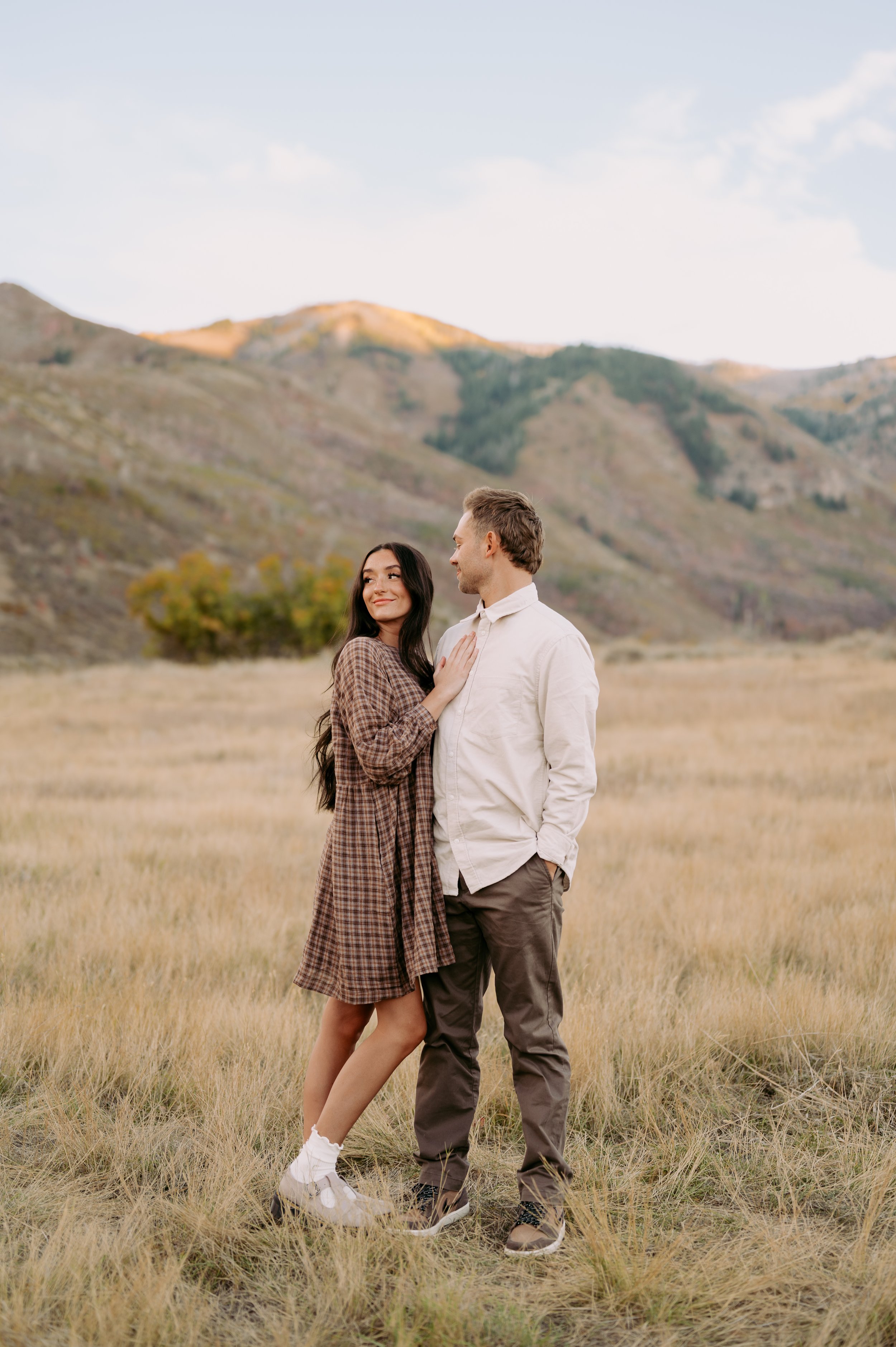 A couple standing in a grassy field with mountains in the background, the woman resting her hand on the man's chest and smiling at him.
