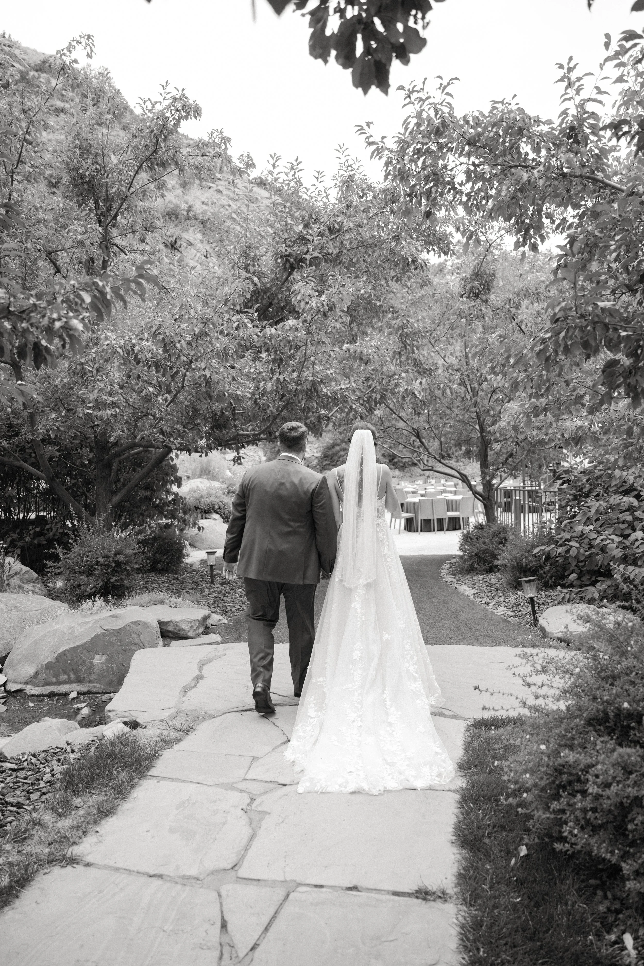 A bride and groom walking on a stone path outdoors, surrounded by trees and bushes, with a table set for an event in the background.