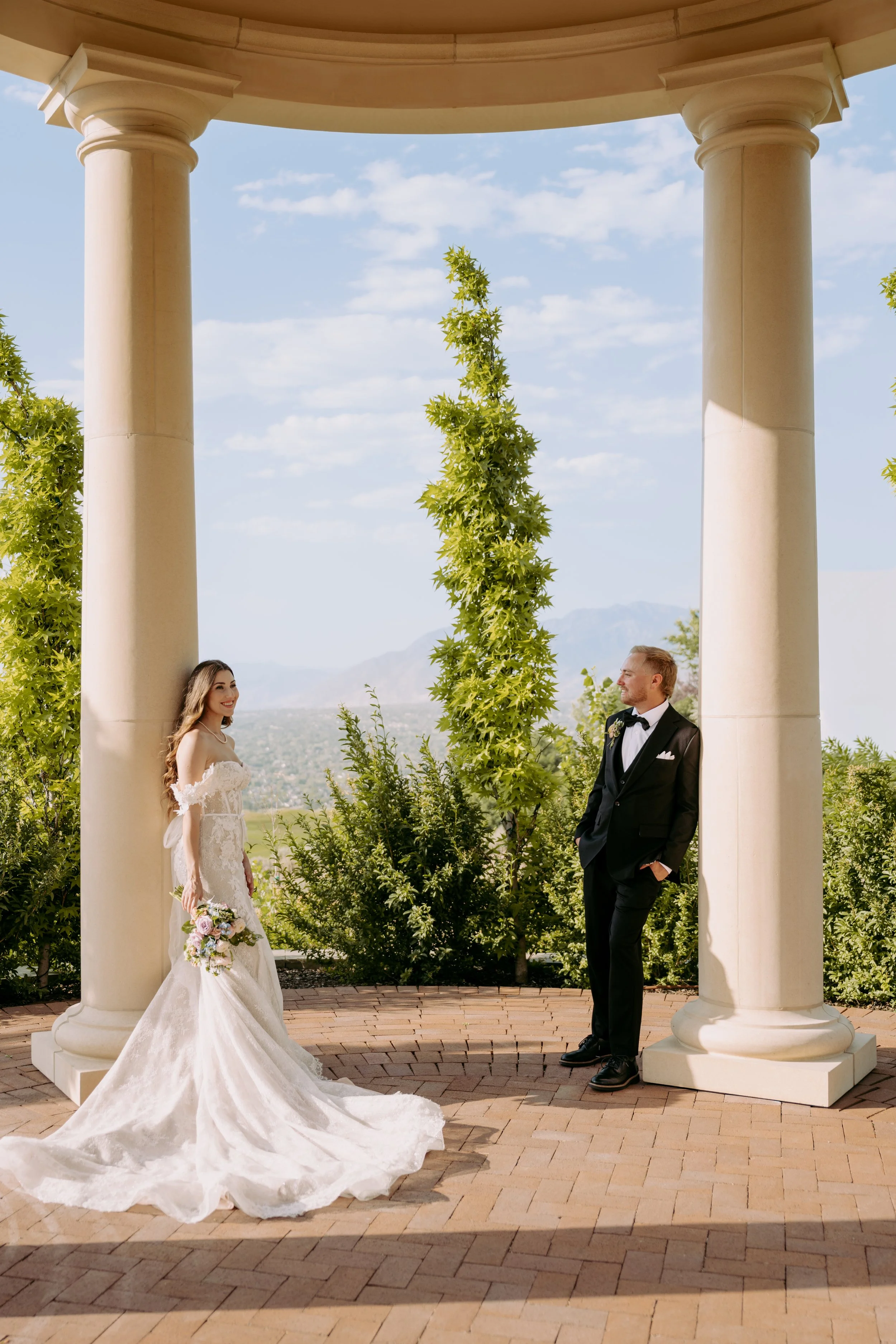 A bride in a white wedding dress holding a bouquet and a groom in a black tuxedo with a bow tie standing under a circular pavilion with large columns, outdoor setting with trees and mountains in the background on a sunny day.