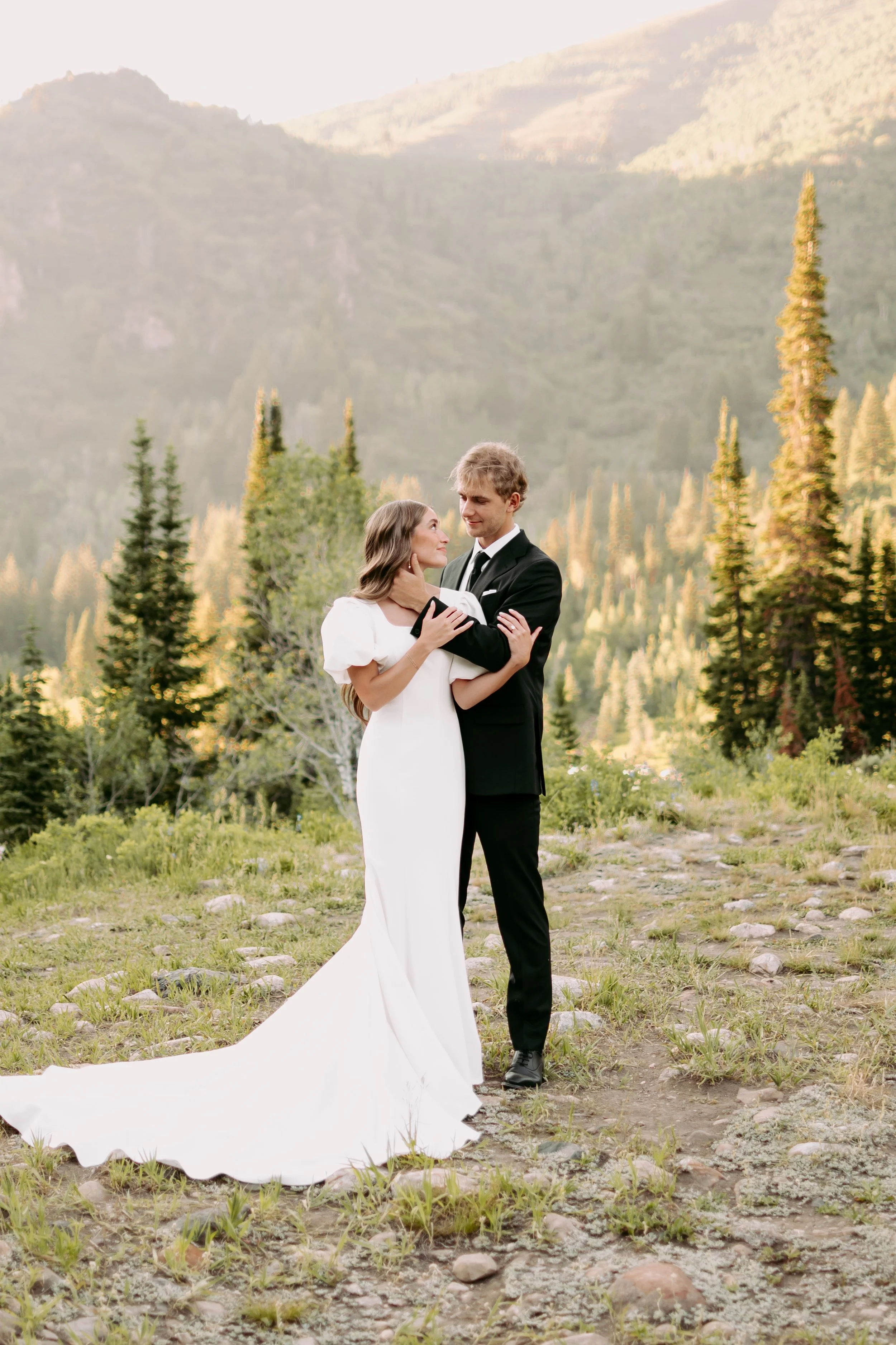 A bride and groom embrace in a scenic outdoor setting with forested mountains in the background. The bride wears a white gown with a long train, and the groom is dressed in a black suit and tie. They gaze into each other's eyes amid a natural landscape with trees and rocky ground.