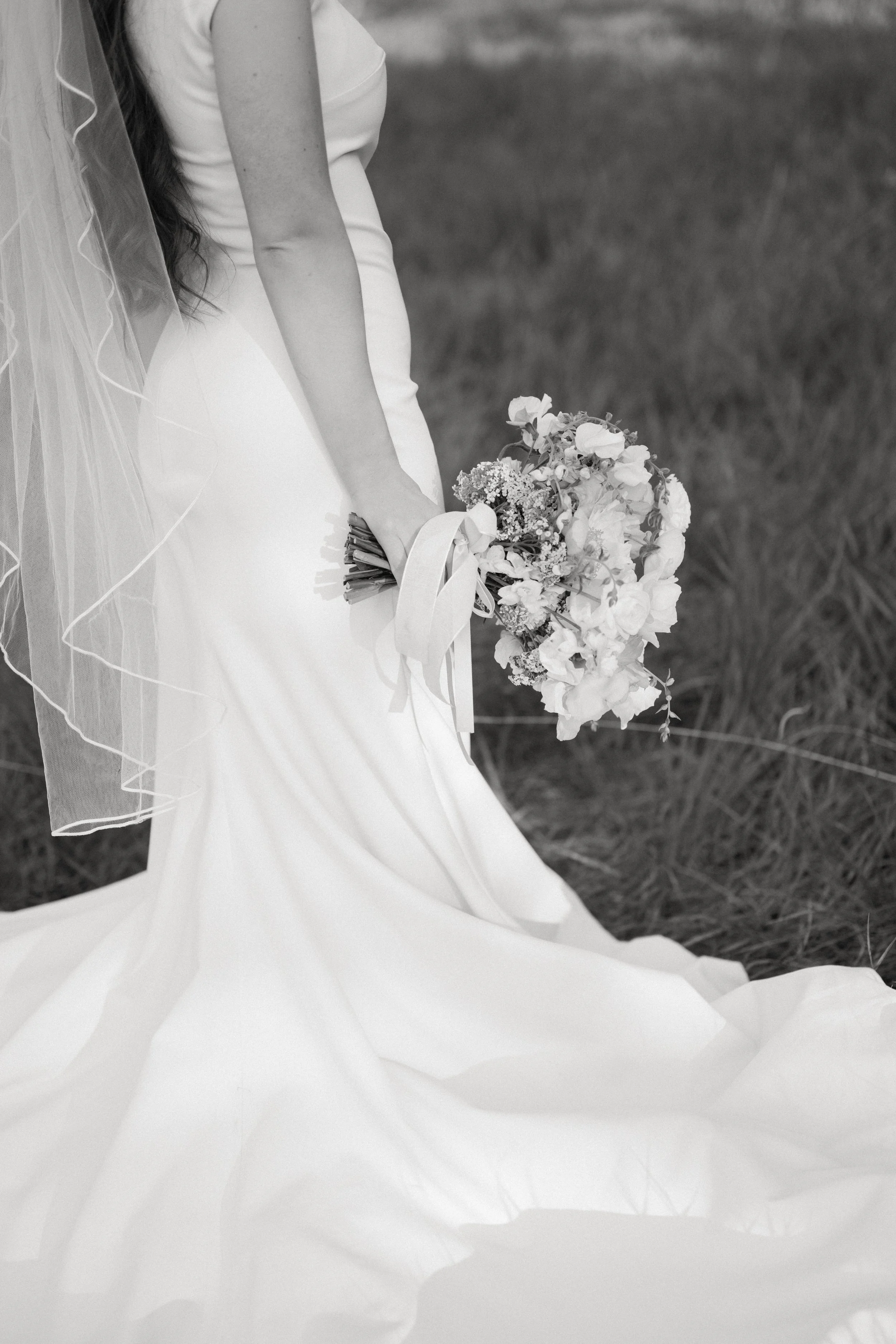 A bride in a white wedding gown holding a bouquet of flowers, standing outdoors on grass.