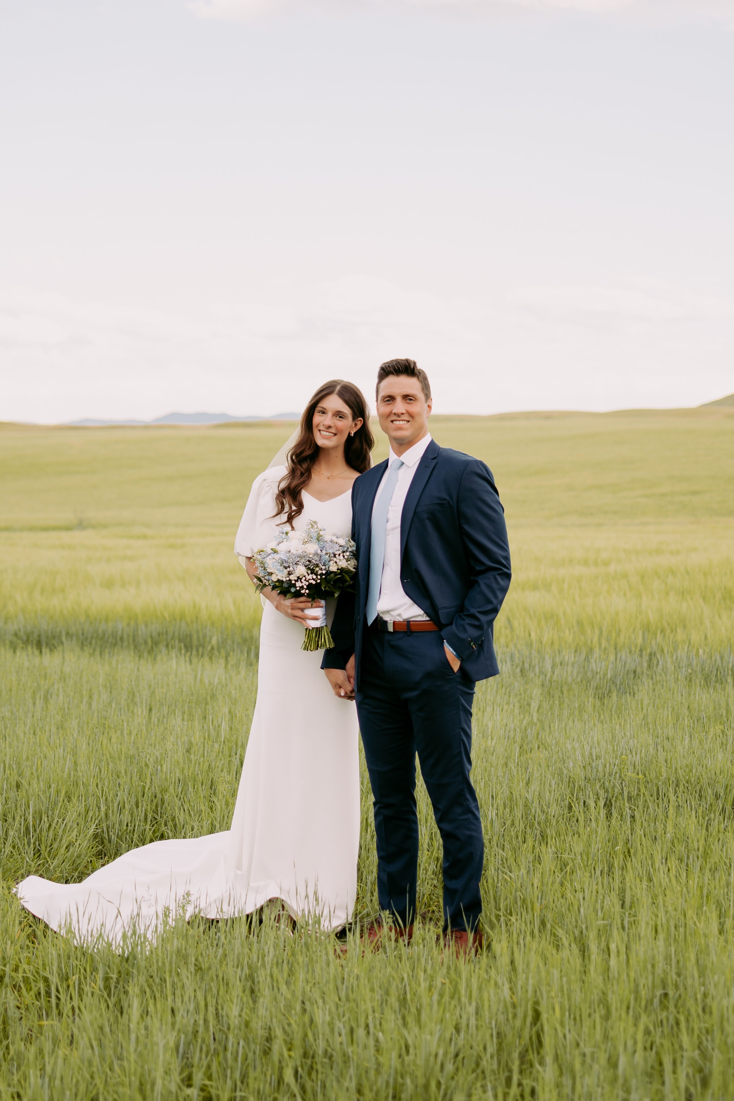 A bride and groom standing hand in hand in a green field with a cloudy sky.