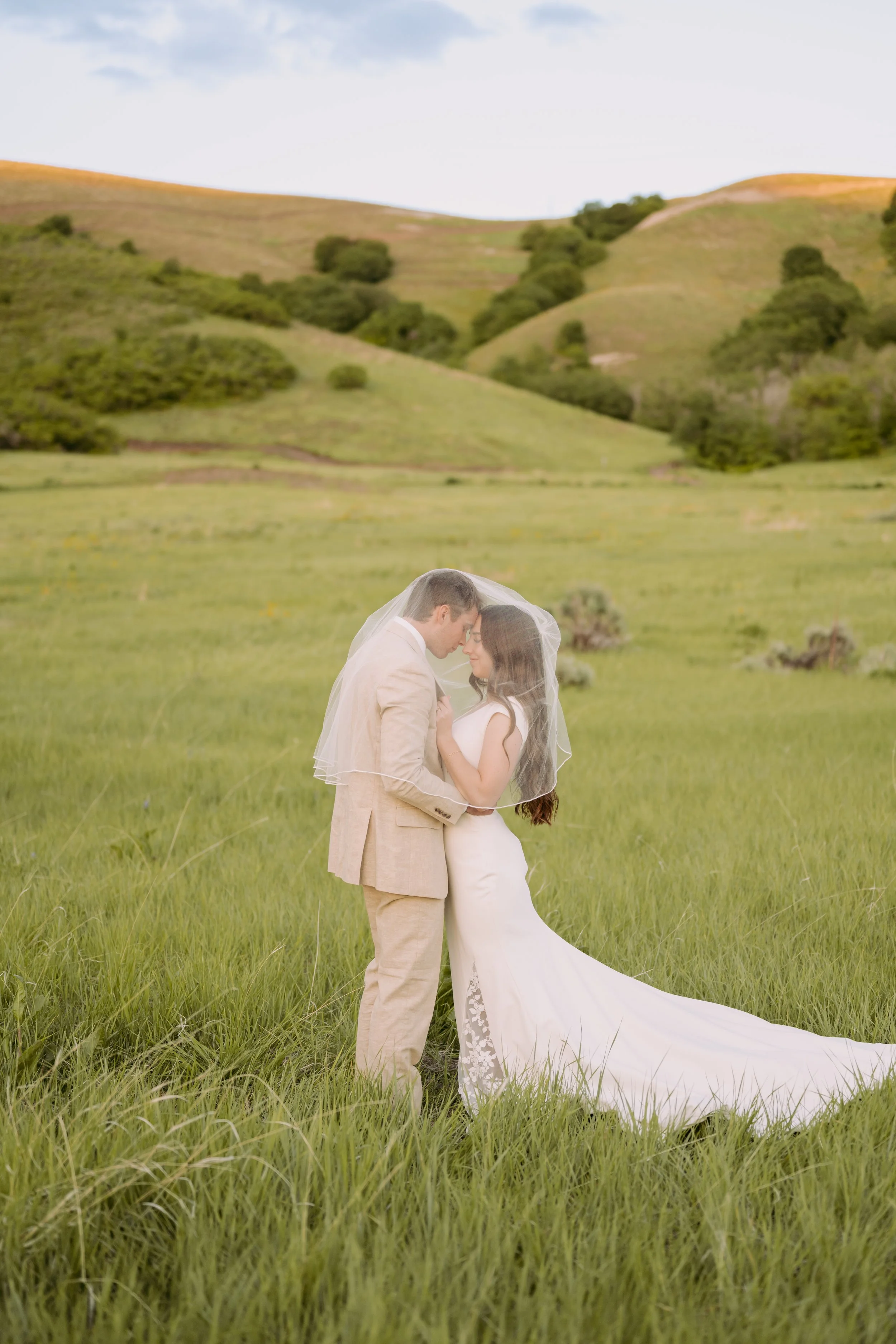 A bride and groom embracing in a grassy field, with hills in the background, on their wedding day.