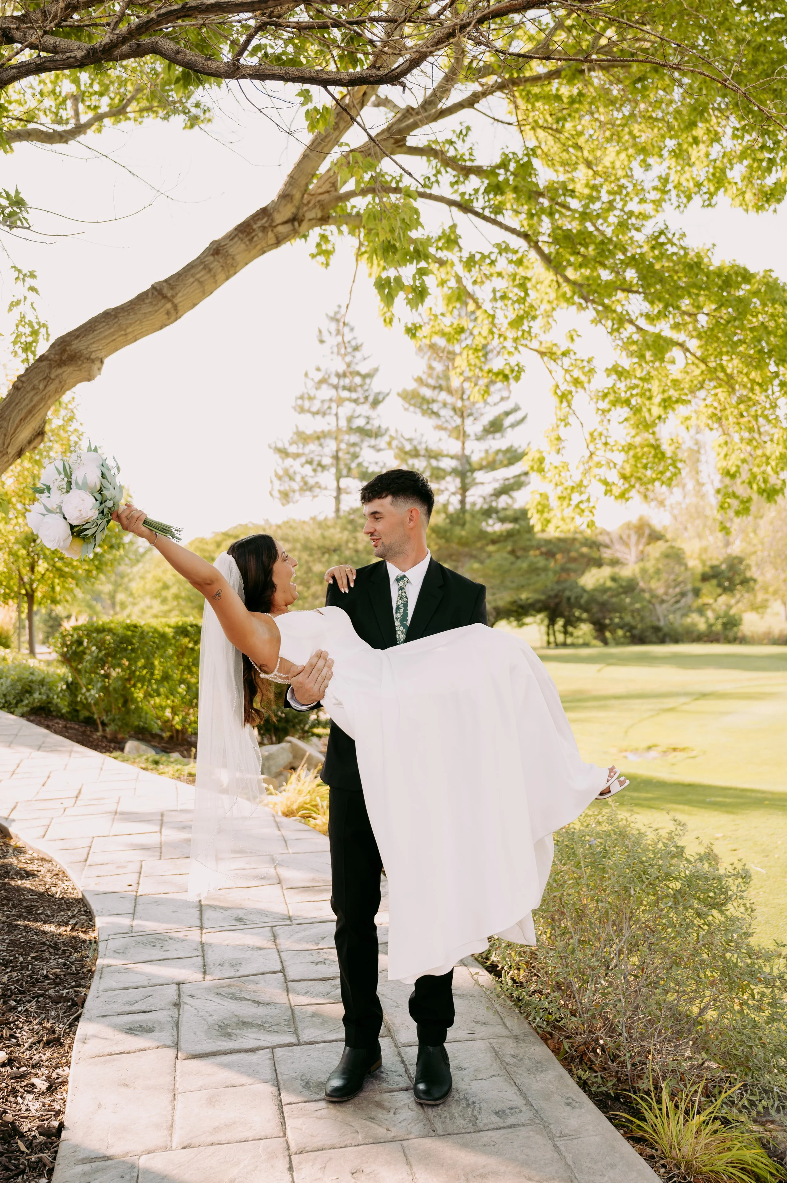 A man in a black suit holding a woman in a white wedding dress with a bouquet of flowers, walking outdoors on a stone path surrounded by greenery and trees.