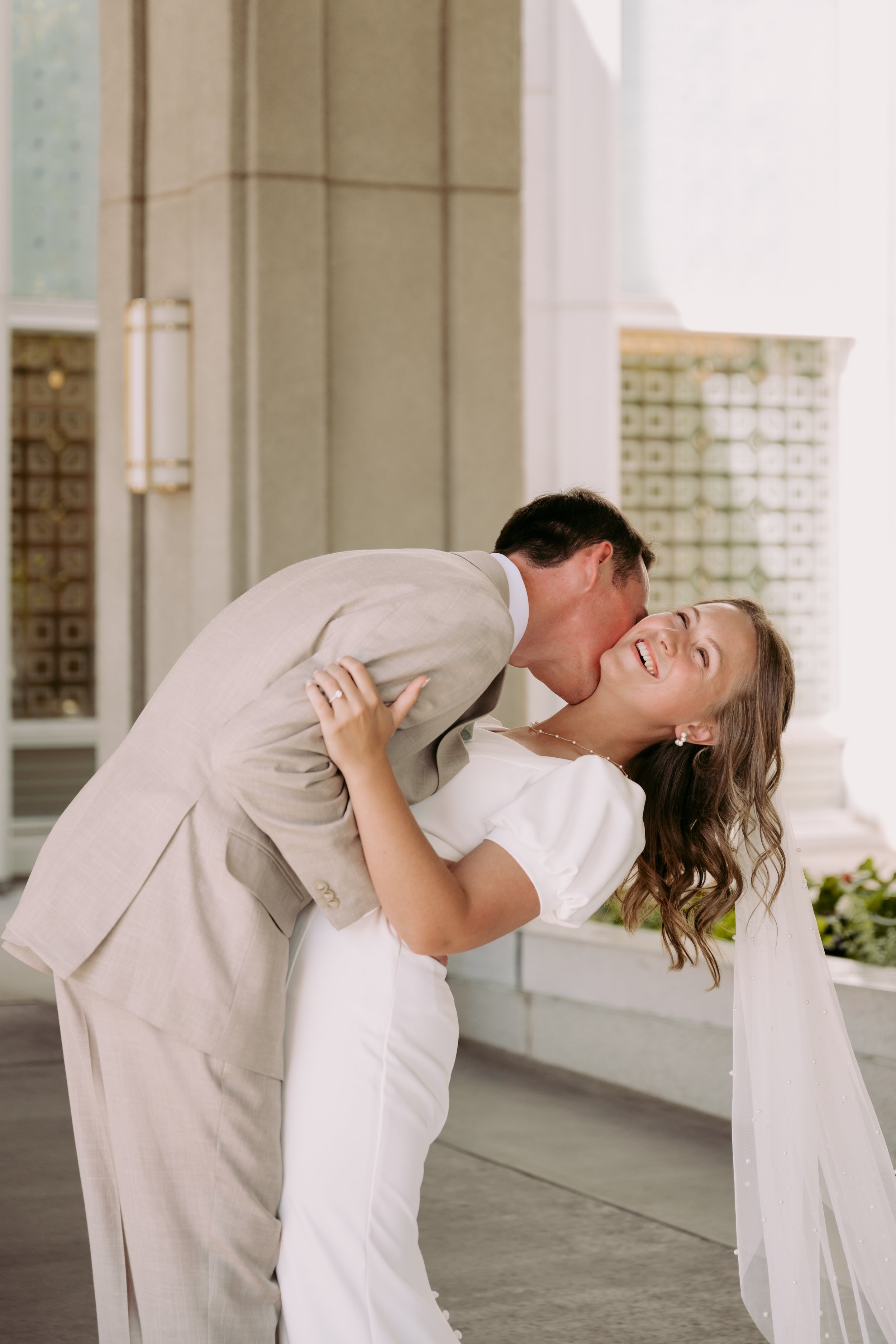 A couple on their wedding day, with the groom kissing the bride on her cheek as she smiles happily, in a bright, elegant setting.