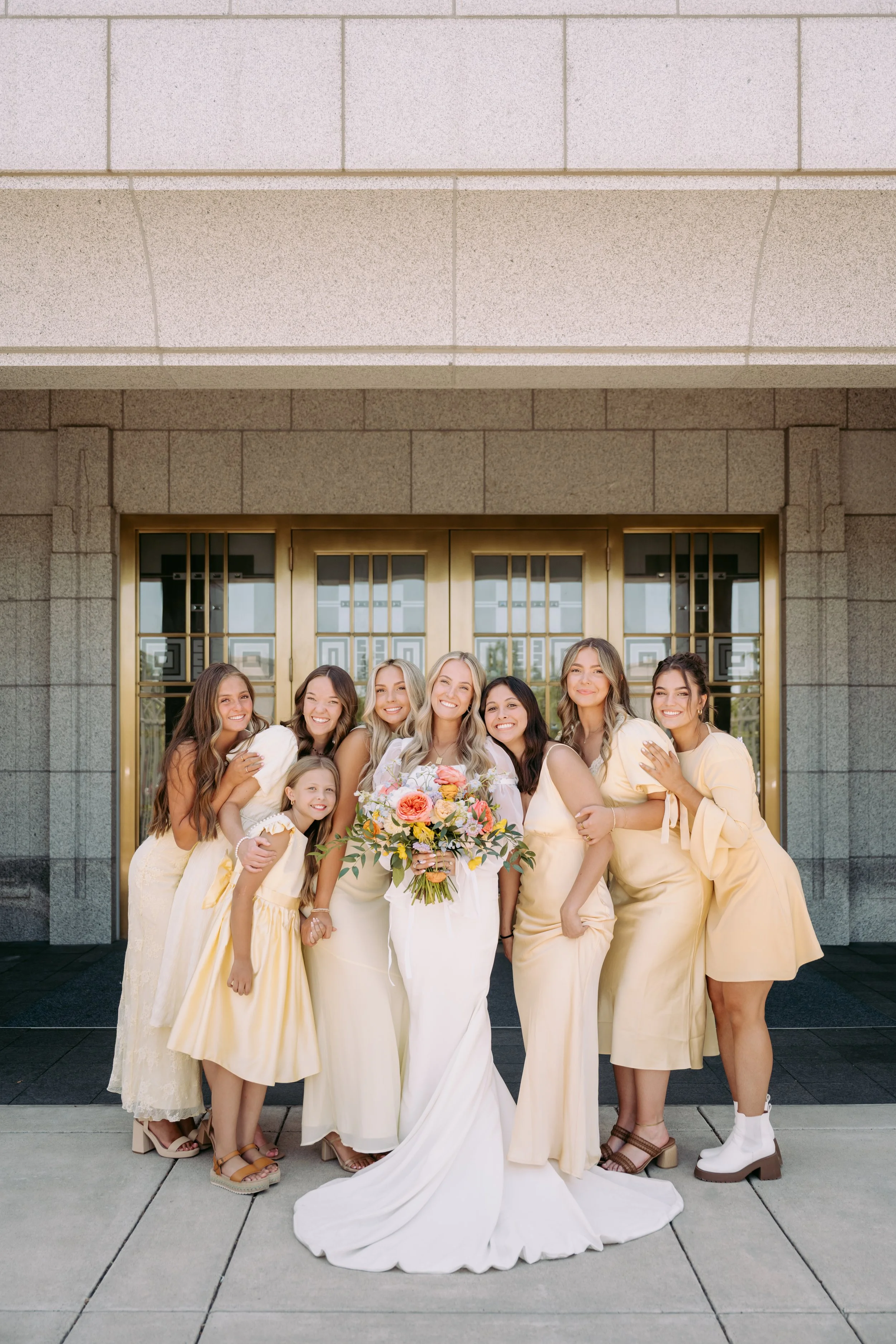 A group of eight women, including a bride with a bouquet, standing outside a building with a large glass doorway, all dressed in light-colored dresses.