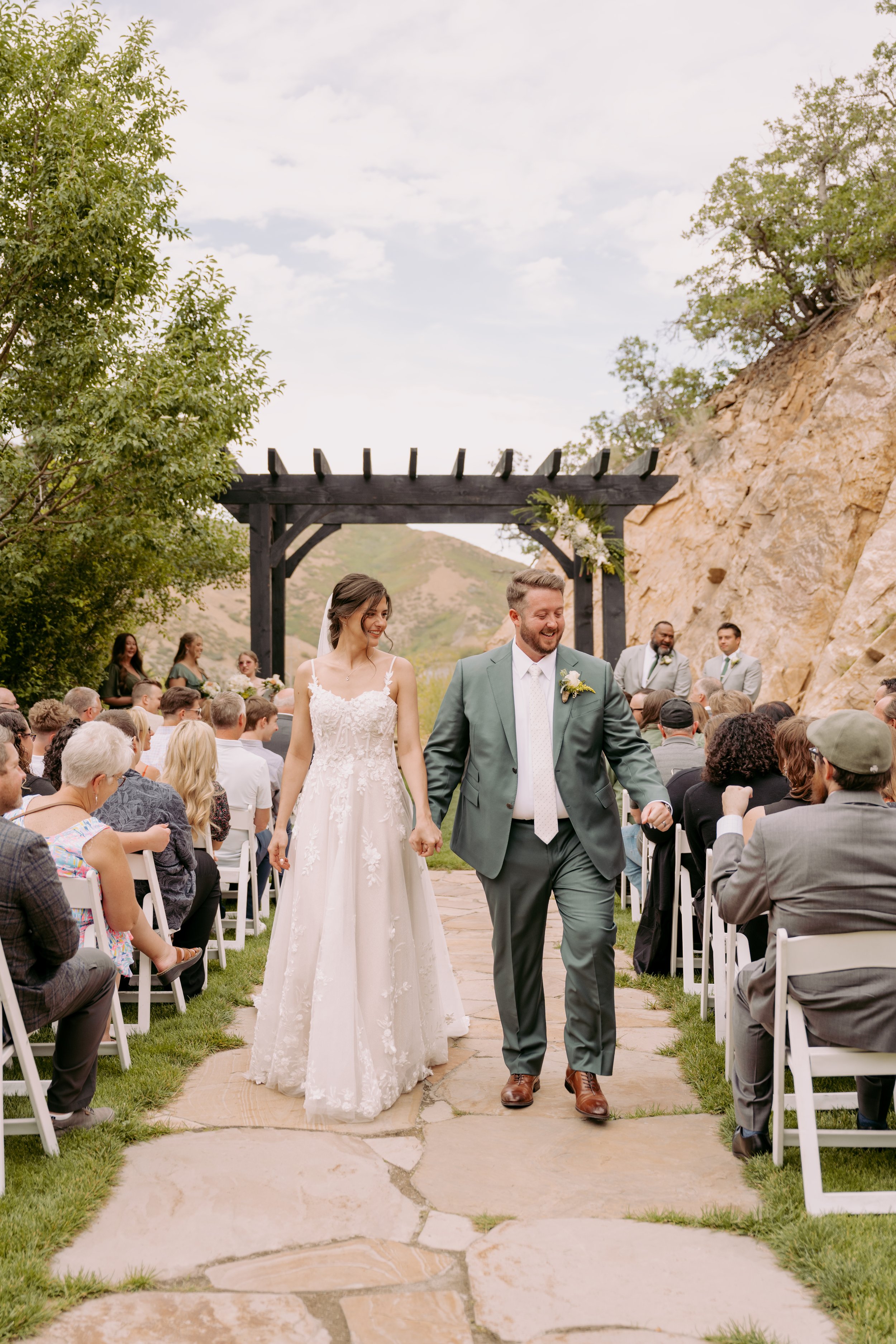 A newly married couple walking hand in hand down the aisle at an outdoor wedding ceremony set against a scenic mountainous background. The bride is wearing a white lace wedding dress, and the groom is dressed in a green suit with a boutonniere.