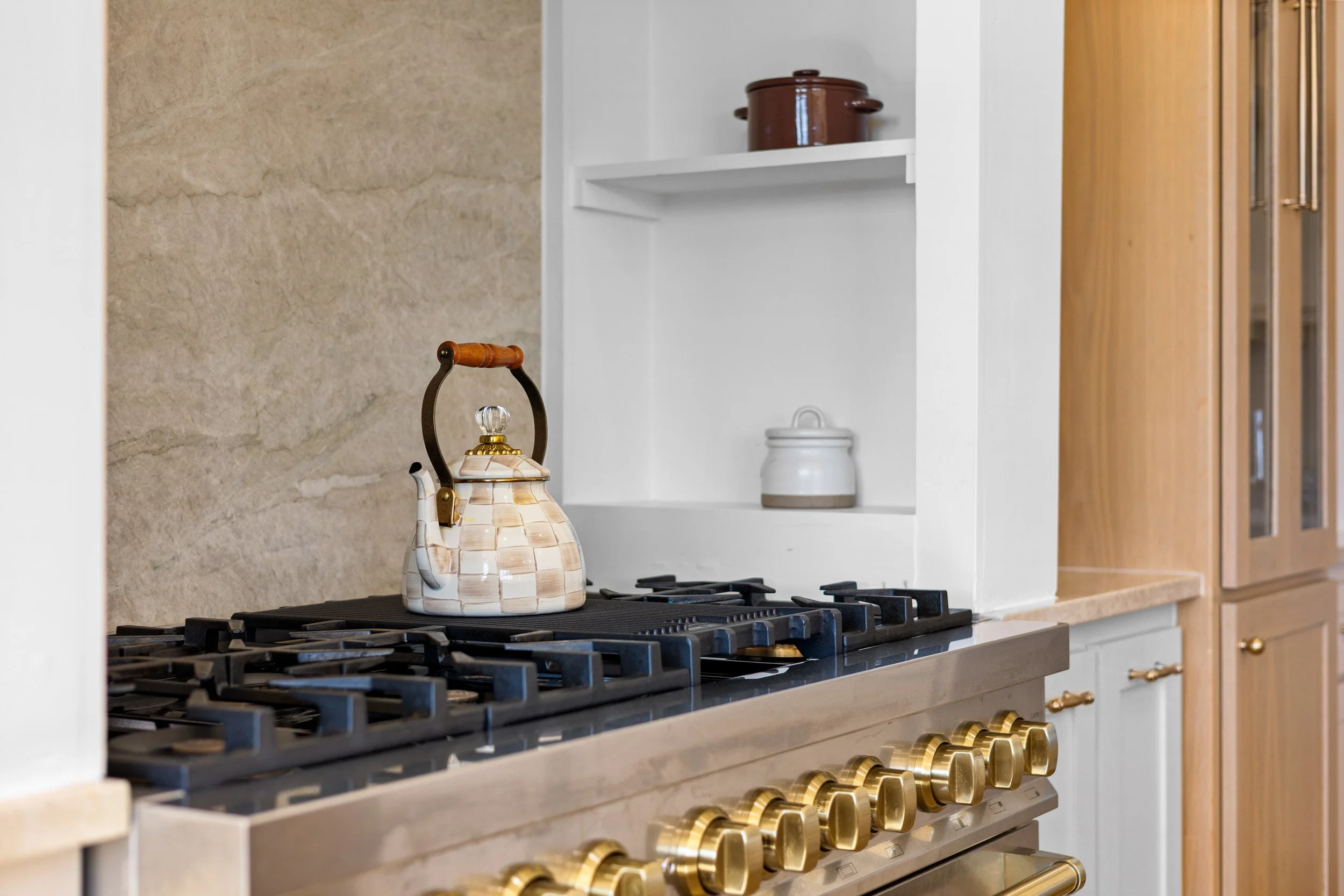 Kitchen stove with a kettle on top, beige stone backsplash, white and wood cabinetry, and shelves with cookware.