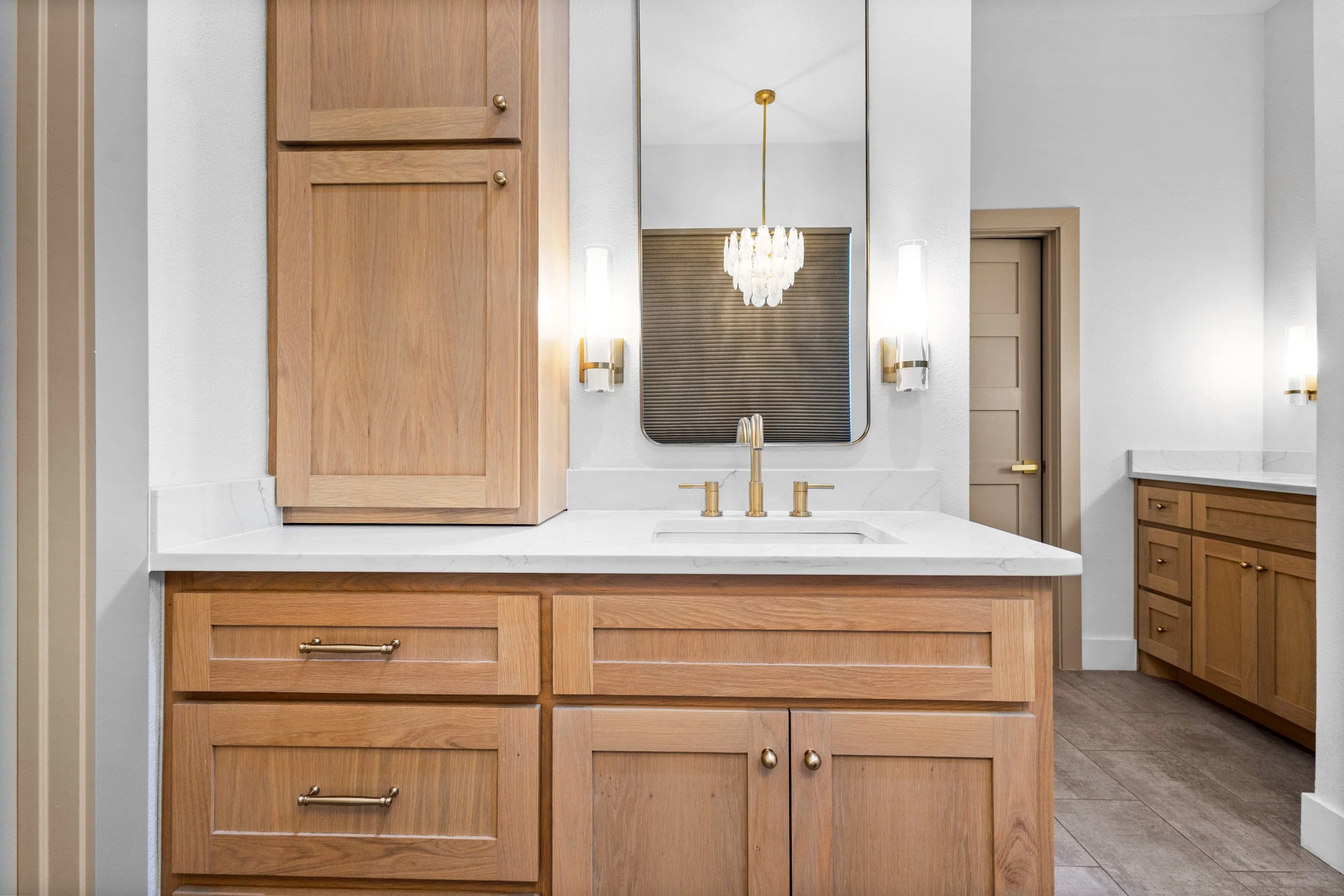 A modern bathroom vanity area with wooden cabinets, a white countertop, gold hardware, and a large mirror. There are two wall-mounted lights on either side of the mirror, and a chandelier hanging from the ceiling reflected in the mirror. A gray door 
