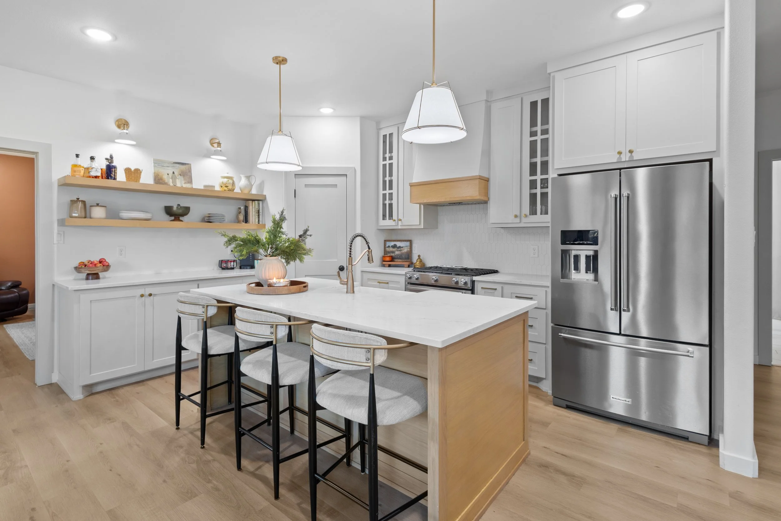 Modern kitchen with white cabinets, stainless steel appliances, a central island with barstools, and wood flooring. Pendant lights hang above the island, and open shelves display dishes and decorative items.