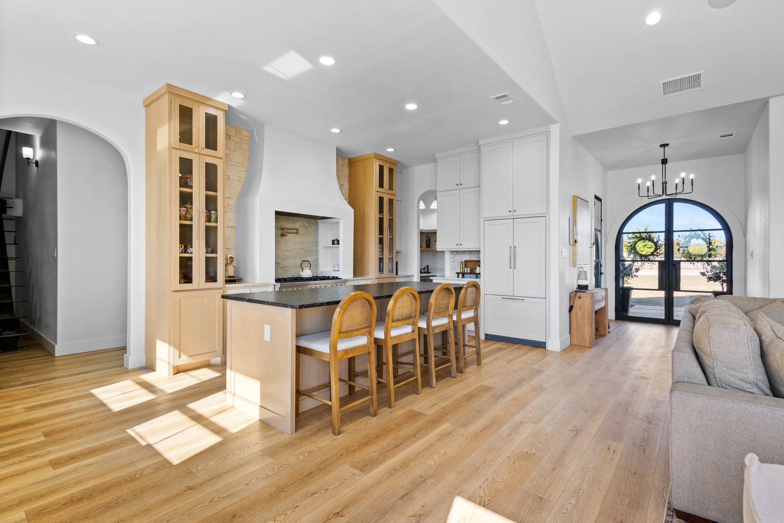 Open-concept kitchen with island, four wooden chairs, white cabinets, and a glass door leading outside, with hardwood floors and a beige sofa in the living area.